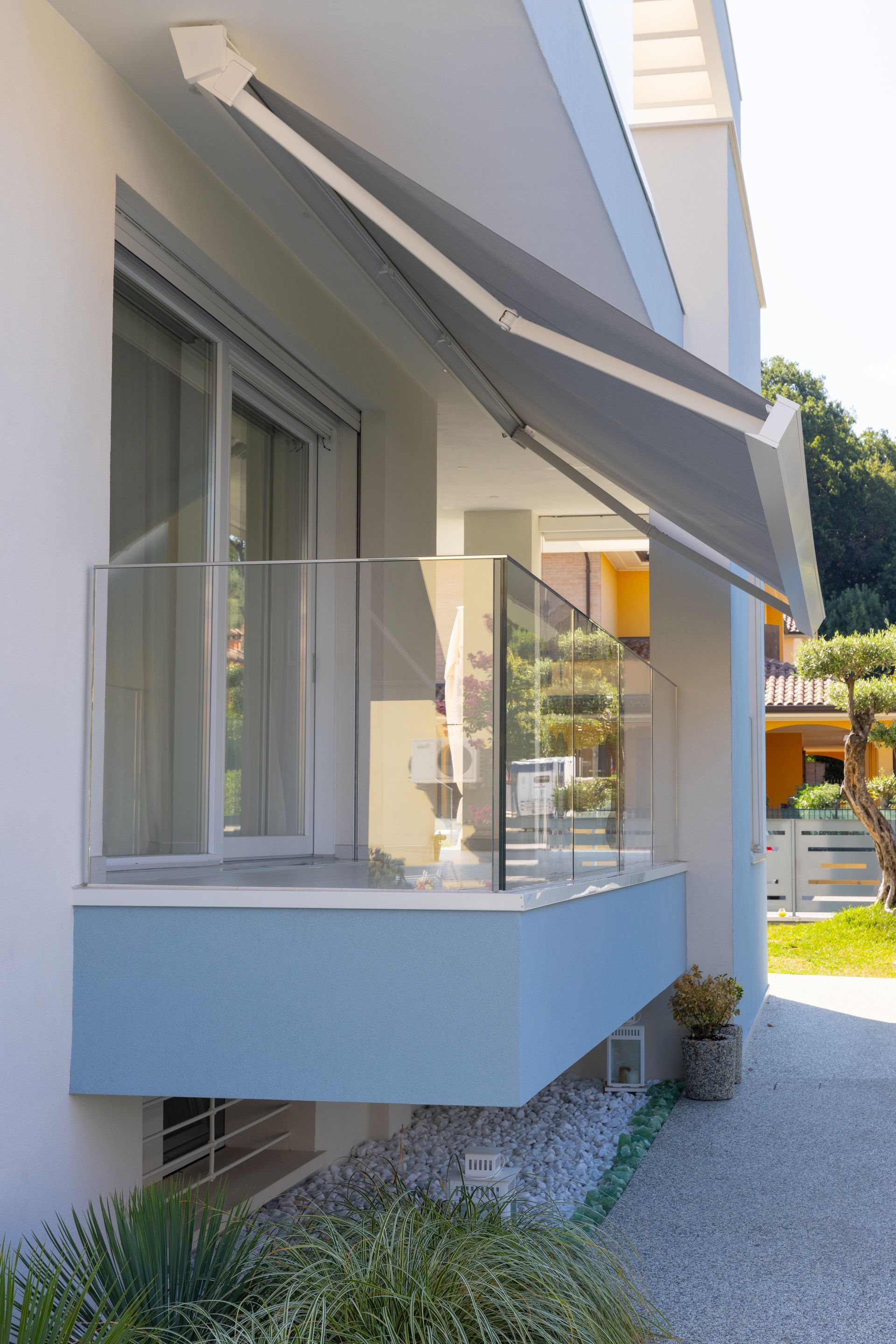 Un balcone al piano terra con ringhiera in vetro e tenda da sole retrattile grigia, annesso a una casa azzurra e bianca.