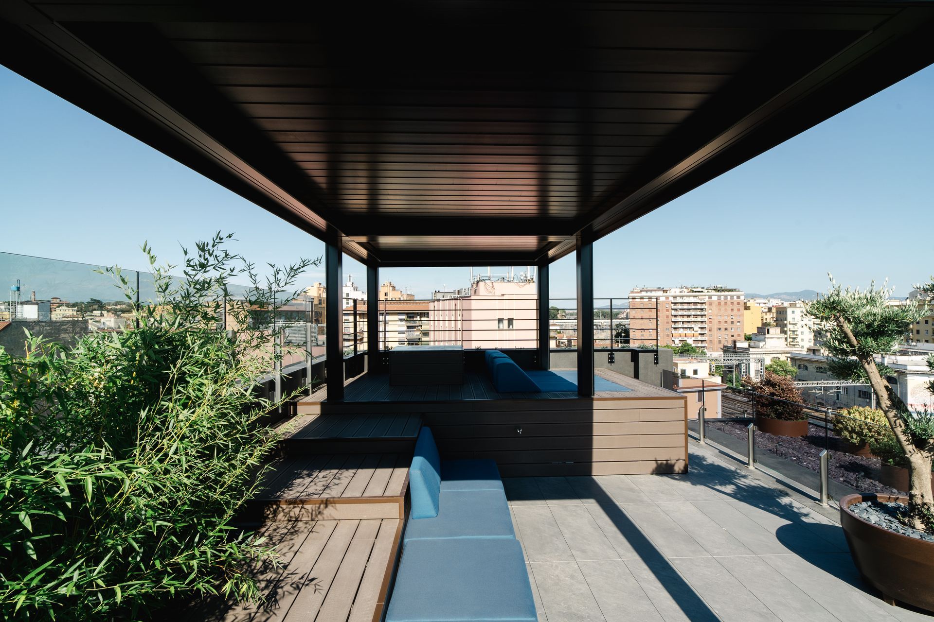 Una terrazza panoramica in legno con una zona relax riparata, arredata con cuscini blu, piante e con vista sulla città.
