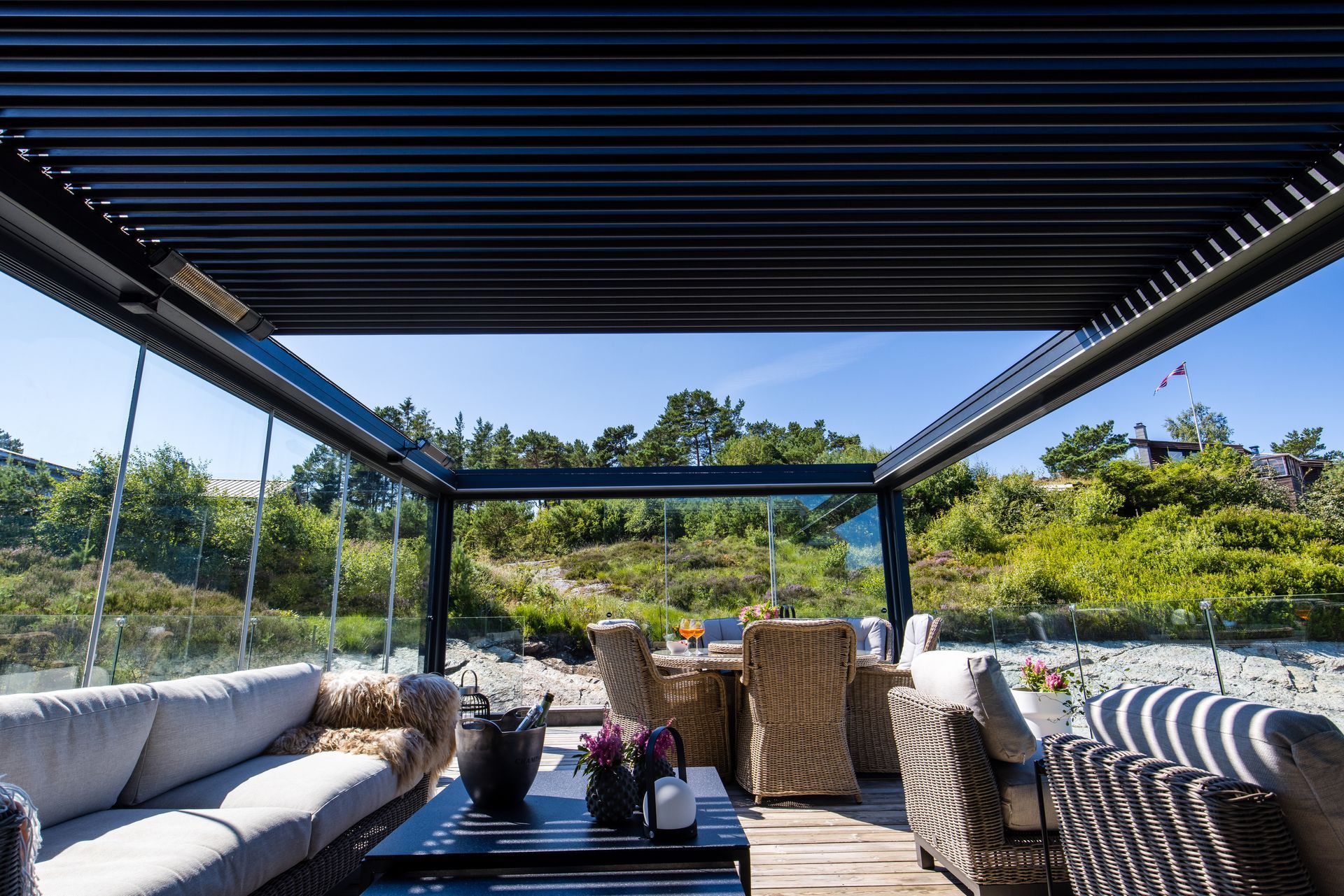 Un moderno patio esterno coperto con divani, un set da pranzo e ringhiere in vetro che si affacciano su un paesaggio soleggiato e erboso.