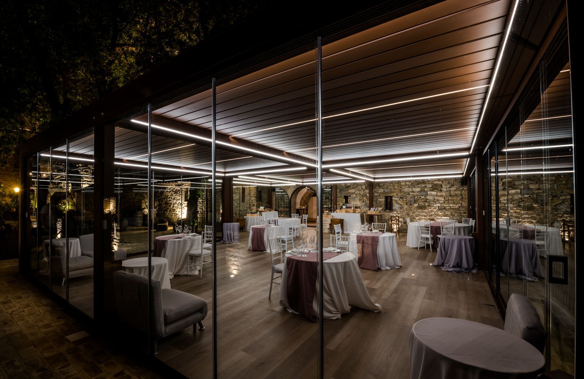 Una struttura da patio illuminata e all'aperto con pareti in vetro, predisposta per eventi serali con tavoli e sedie.