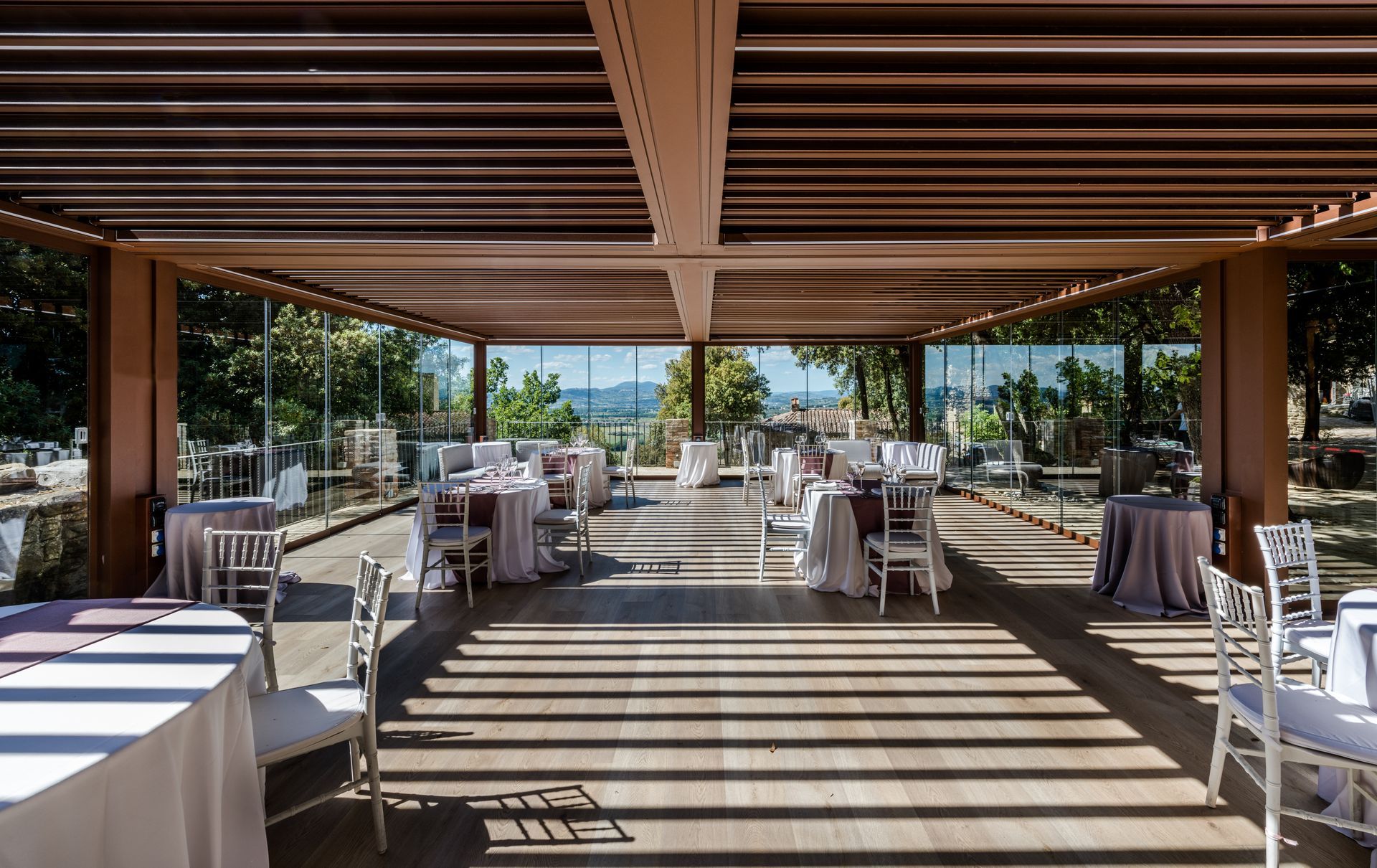 Uno spazio esterno coperto per eventi, con tavoli rotondi, sedie bianche e pareti in vetro che si affacciano su un paesaggio baciato dal sole.