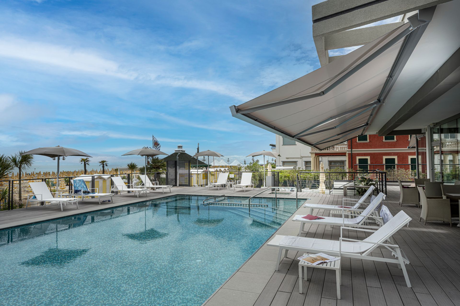 Una piscina turchese circondata da lettini bianchi su una terrazza in legno, sotto un cielo azzurro e limpido.