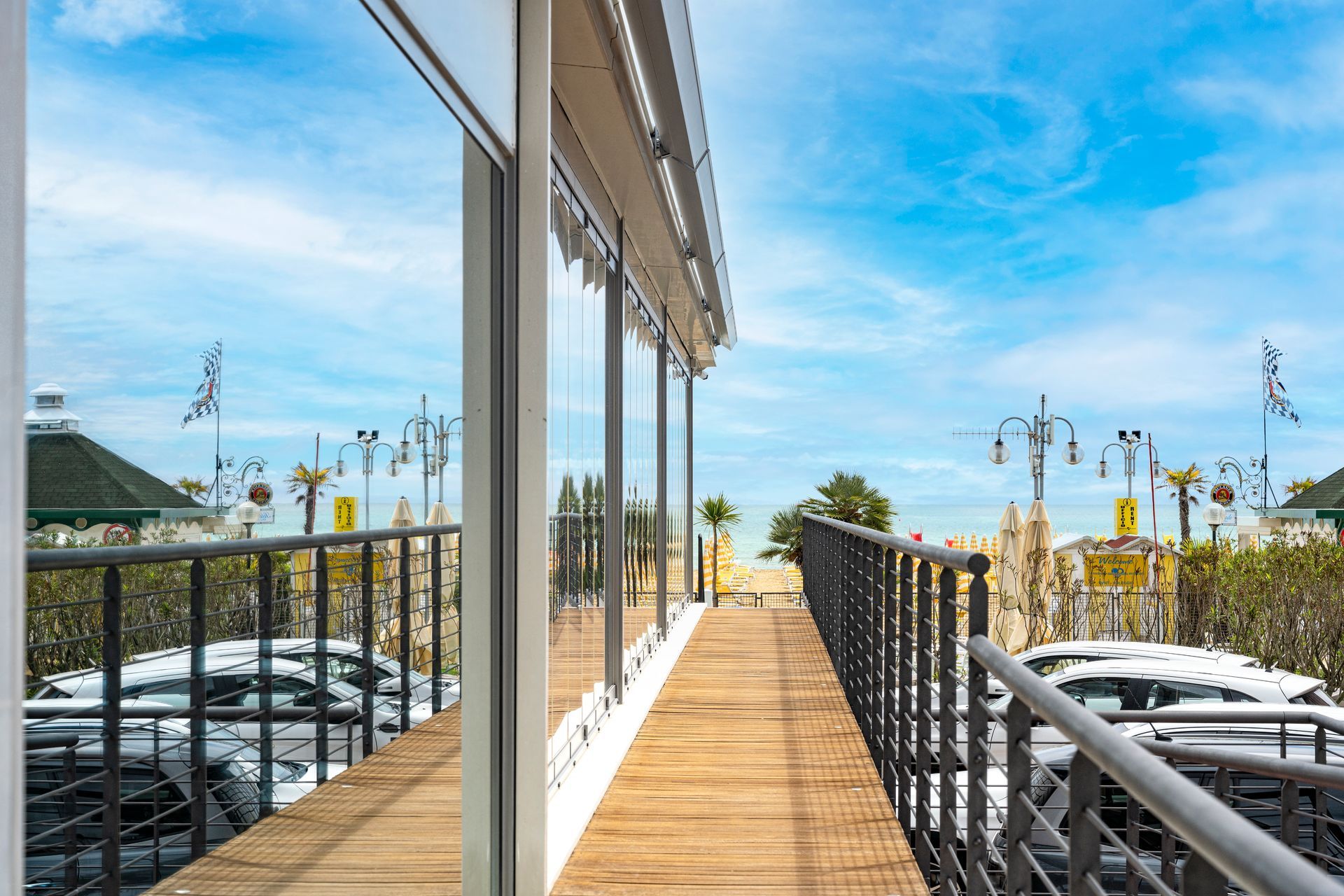 Una passerella di legno si estende tra un moderno edificio in vetro e una ringhiera metallica, affacciandosi su un'area di parcheggio sotto un cielo azzurro.