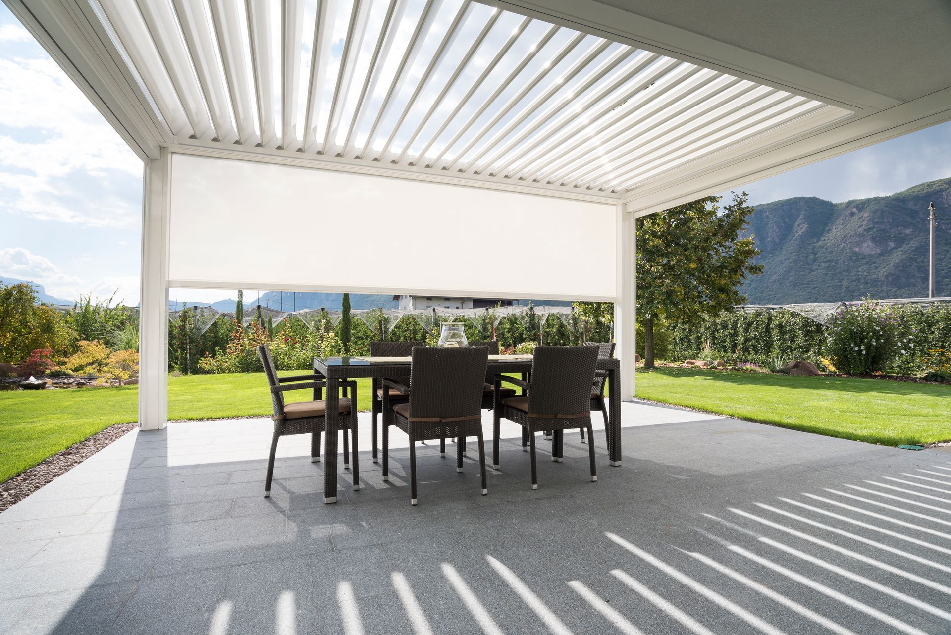 Un set da pranzo da esterno sotto un moderno pergolato con una tenda motorizzata bianca, circondato da un prato e con le montagne sullo sfondo.