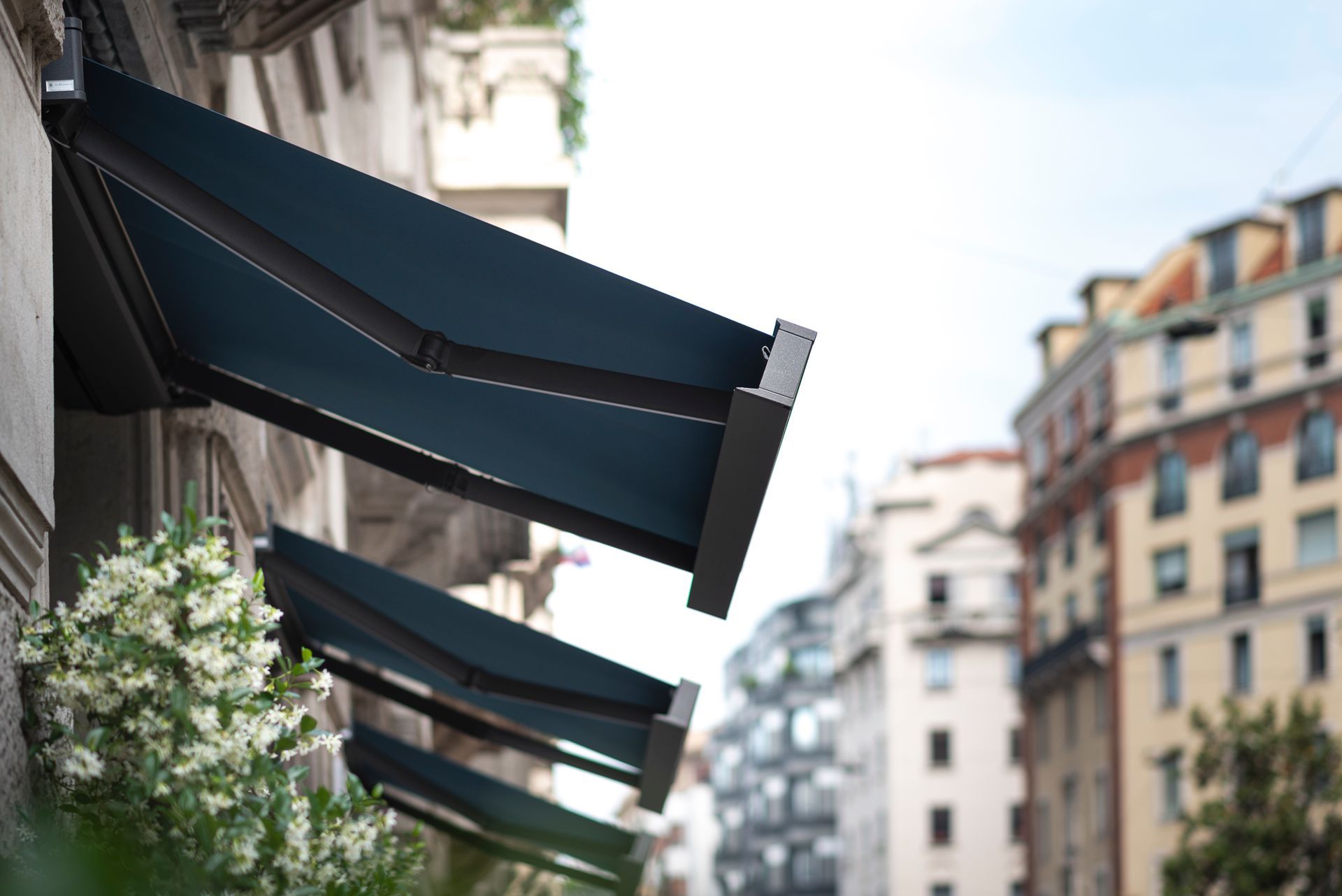 Delle tende da sole inclinate di colore blu scuro si estendono dalla facciata di un edificio, sopra fiori bianchi, affacciandosi su una strada cittadina.