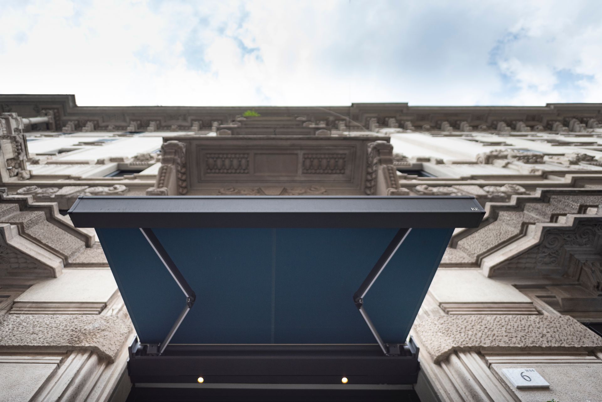 Una moderna tenda da sole blu scuro si estende sopra un ingresso, al di sotto della facciata in pietra di un edificio con dettagli architettonici.