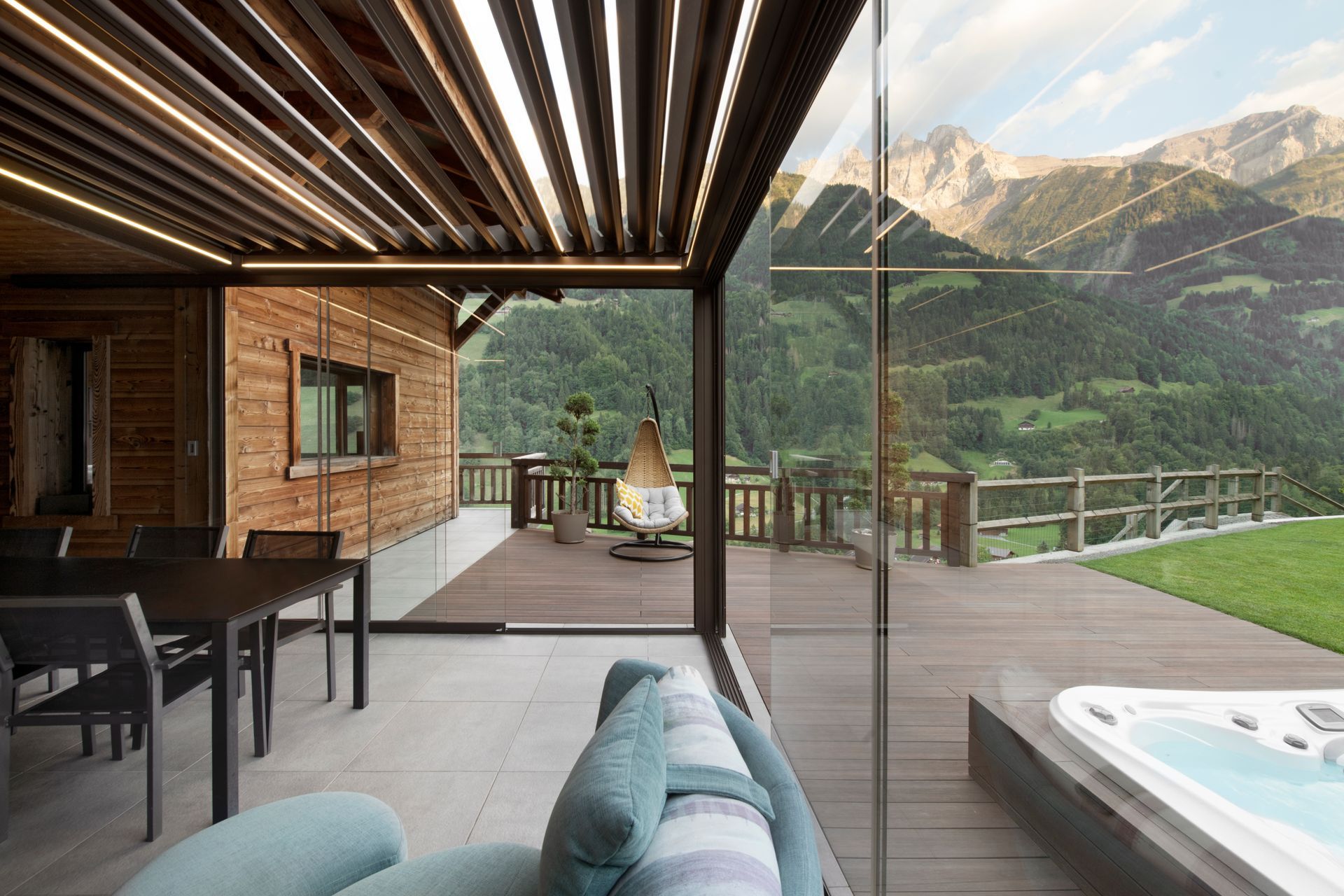 Un moderno patio interno-esterno con set da pranzo, divano e vasca idromassaggio, che si affaccia su una pittoresca valle di montagna al tramonto.