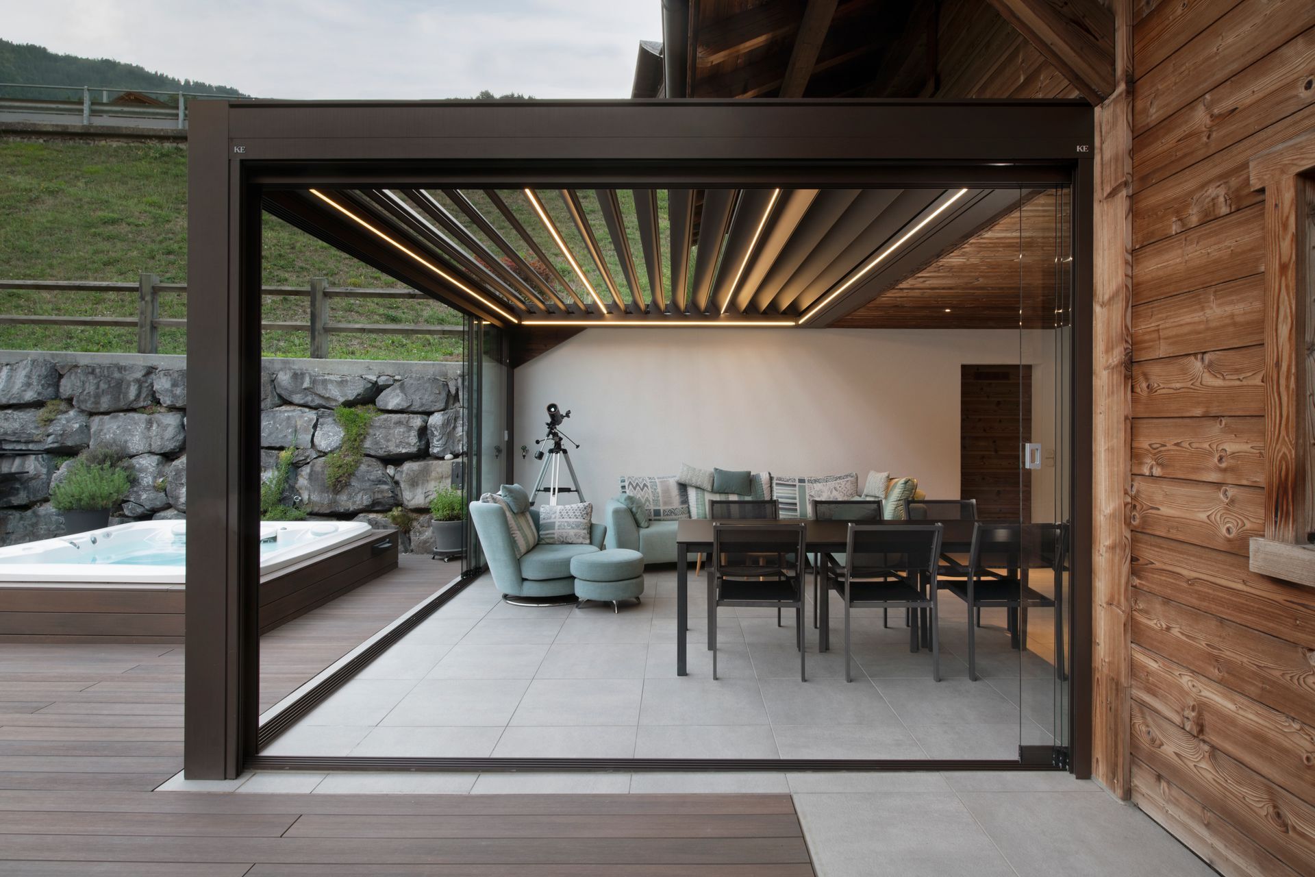 Un moderno patio coperto con illuminazione, mobili da esterno e una vasca idromassaggio accanto a una casetta in legno e un muro in pietra.