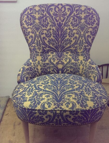 Una poltrona arrotondata con rivestimento in tessuto damascato color crema e viola e gambe in legno chiaro.