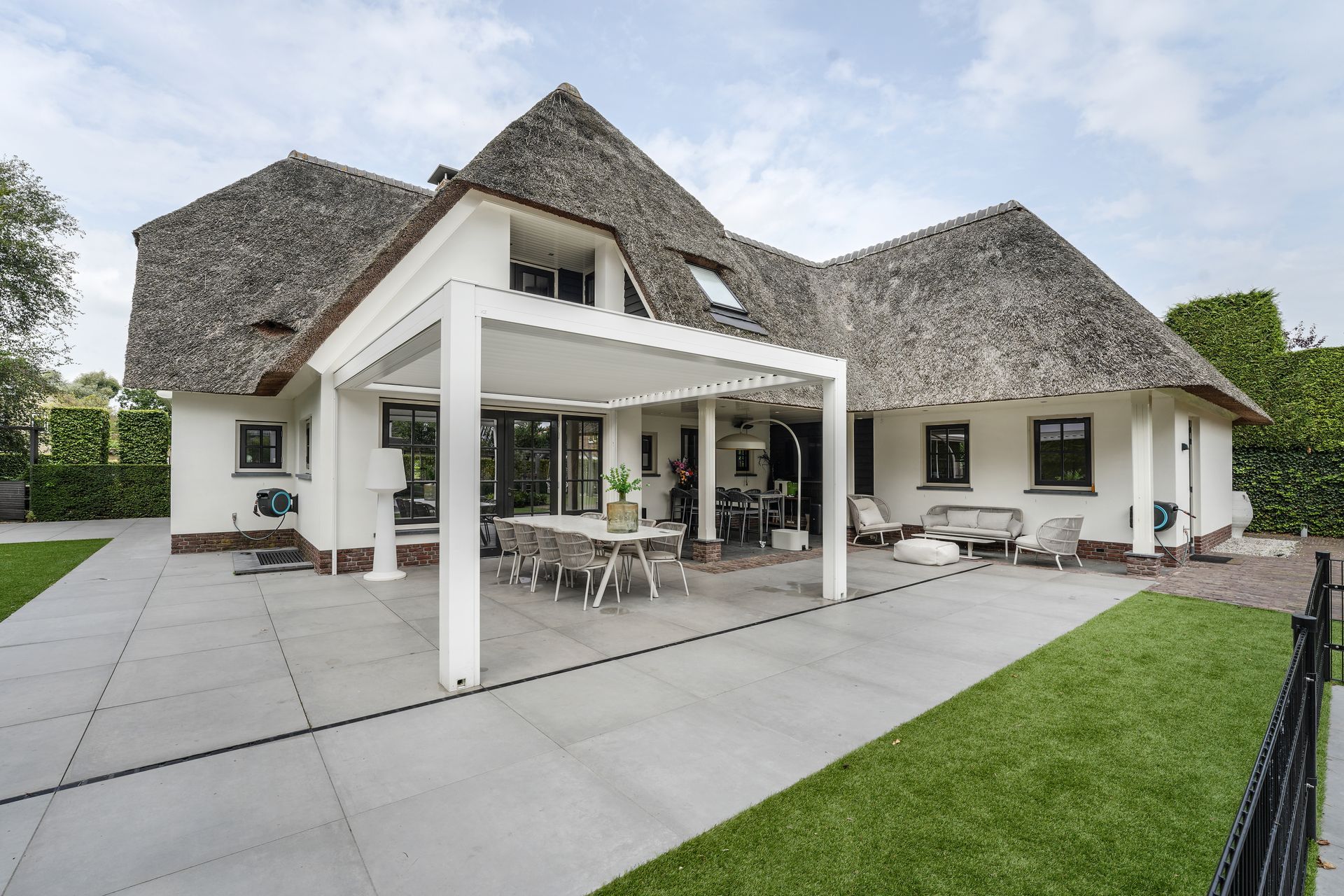 Una casa bianca con tetto di paglia e patio dotato di una moderna zona pranzo coperta, circondata da un prato e da lastre di pavimentazione.