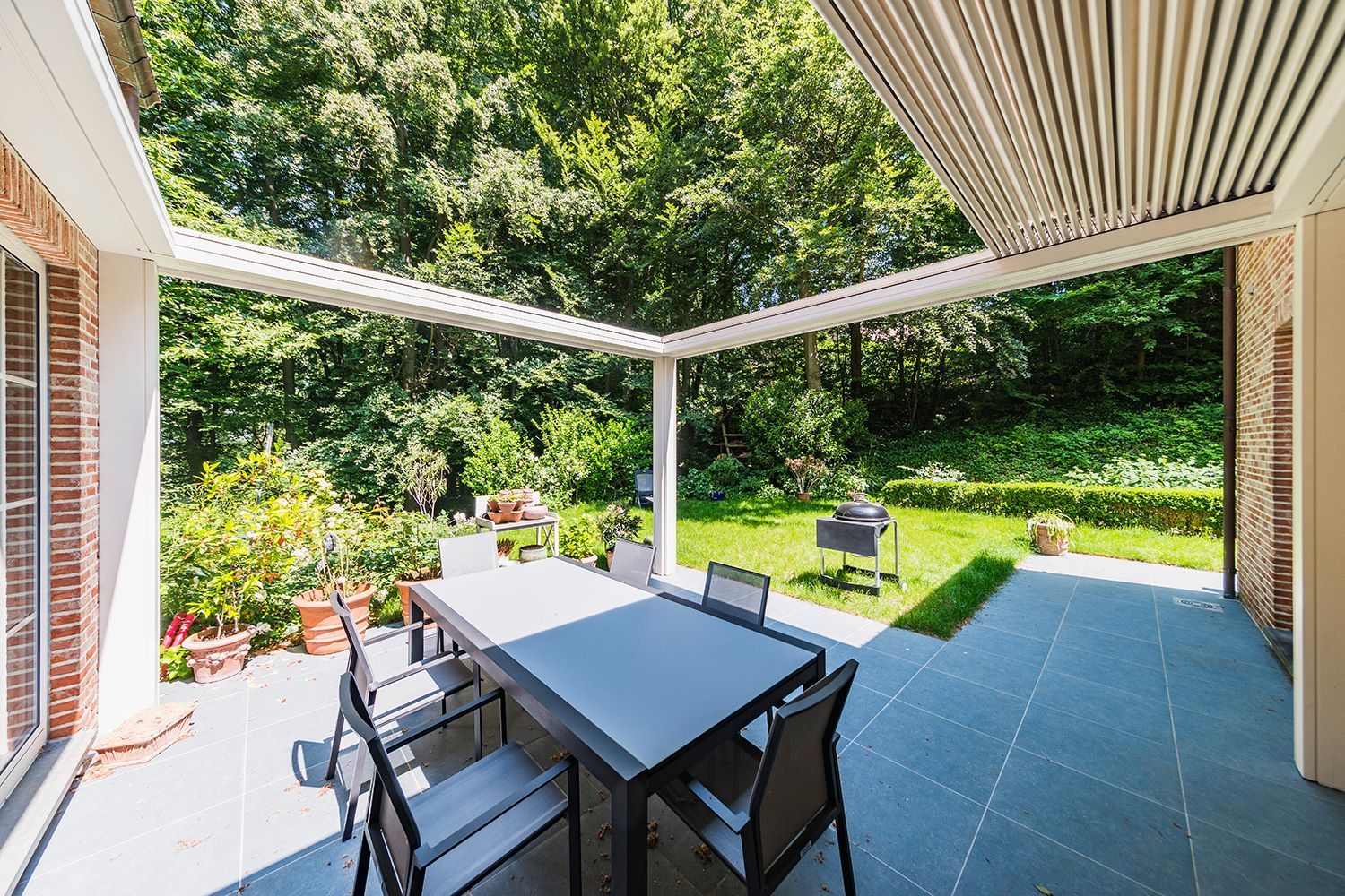 Un set da pranzo da esterno è posizionato sotto un moderno pergolato su una terrazza piastrellata di grigio, con vista su un giardino soleggiato con alberi verdi.