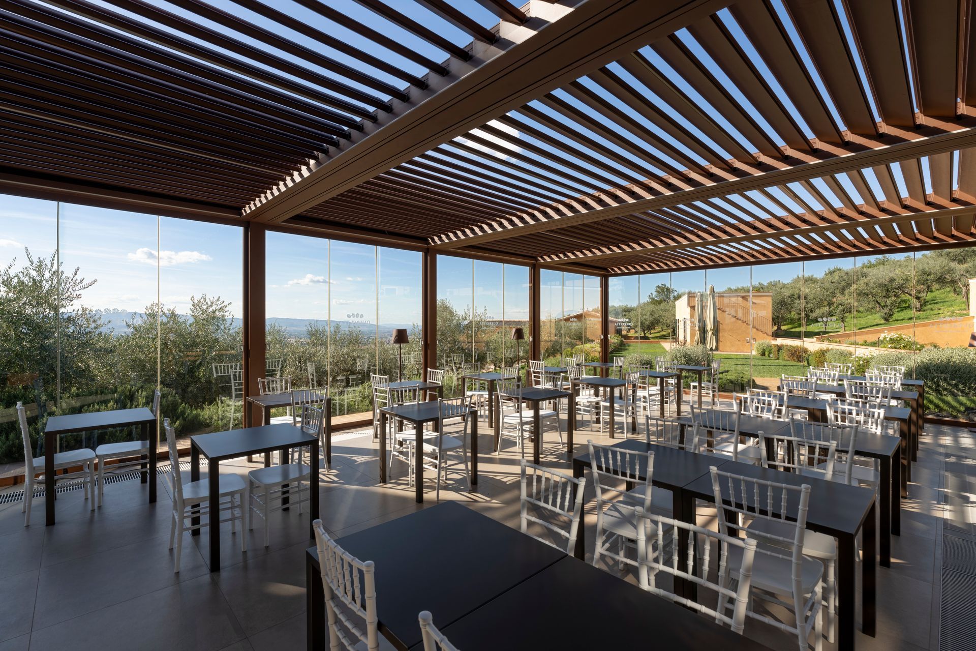 Una terrazza esterna coperta, con file di tavoli e sedie, sotto una tettoia a doghe, che si affaccia su un paesaggio soleggiato.