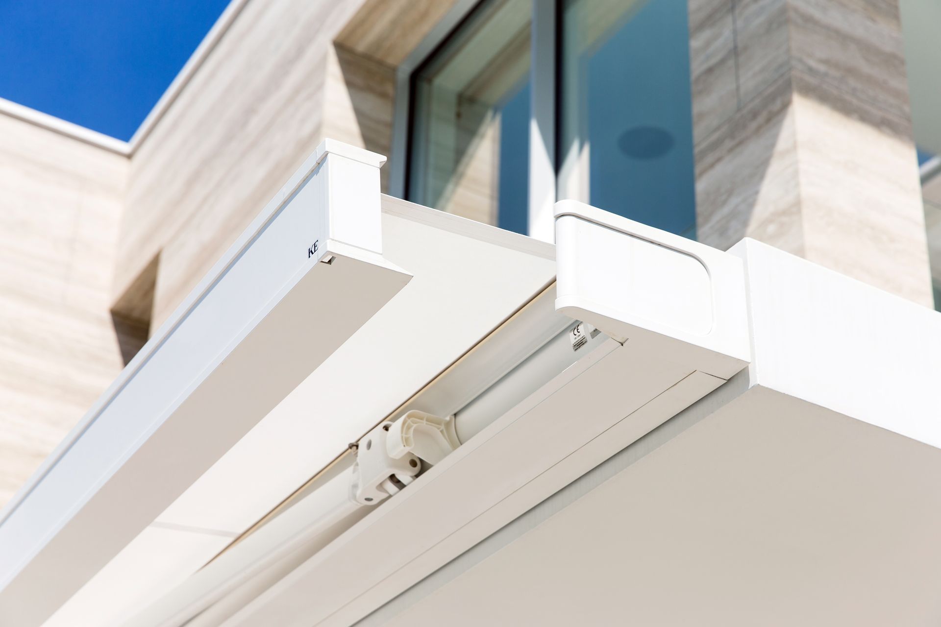 Vista dal basso di una tenda da sole retrattile bianca montata sulla facciata di un edificio moderno, con uno sfondo di cielo azzurro.