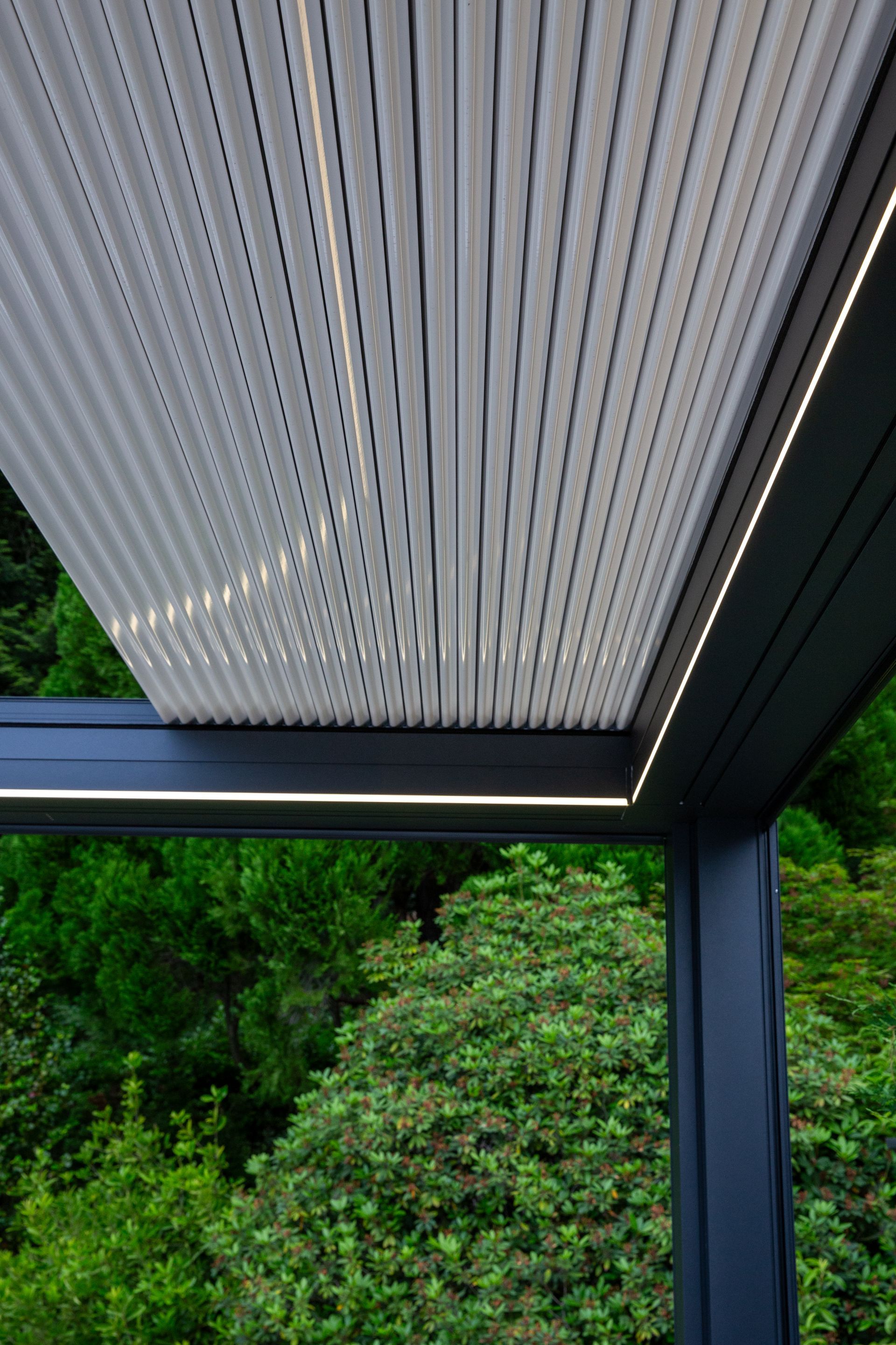 Una pergola bianca retrattile con illuminazione a strisce integrata, parzialmente estesa su un rigoglioso giardino verde.