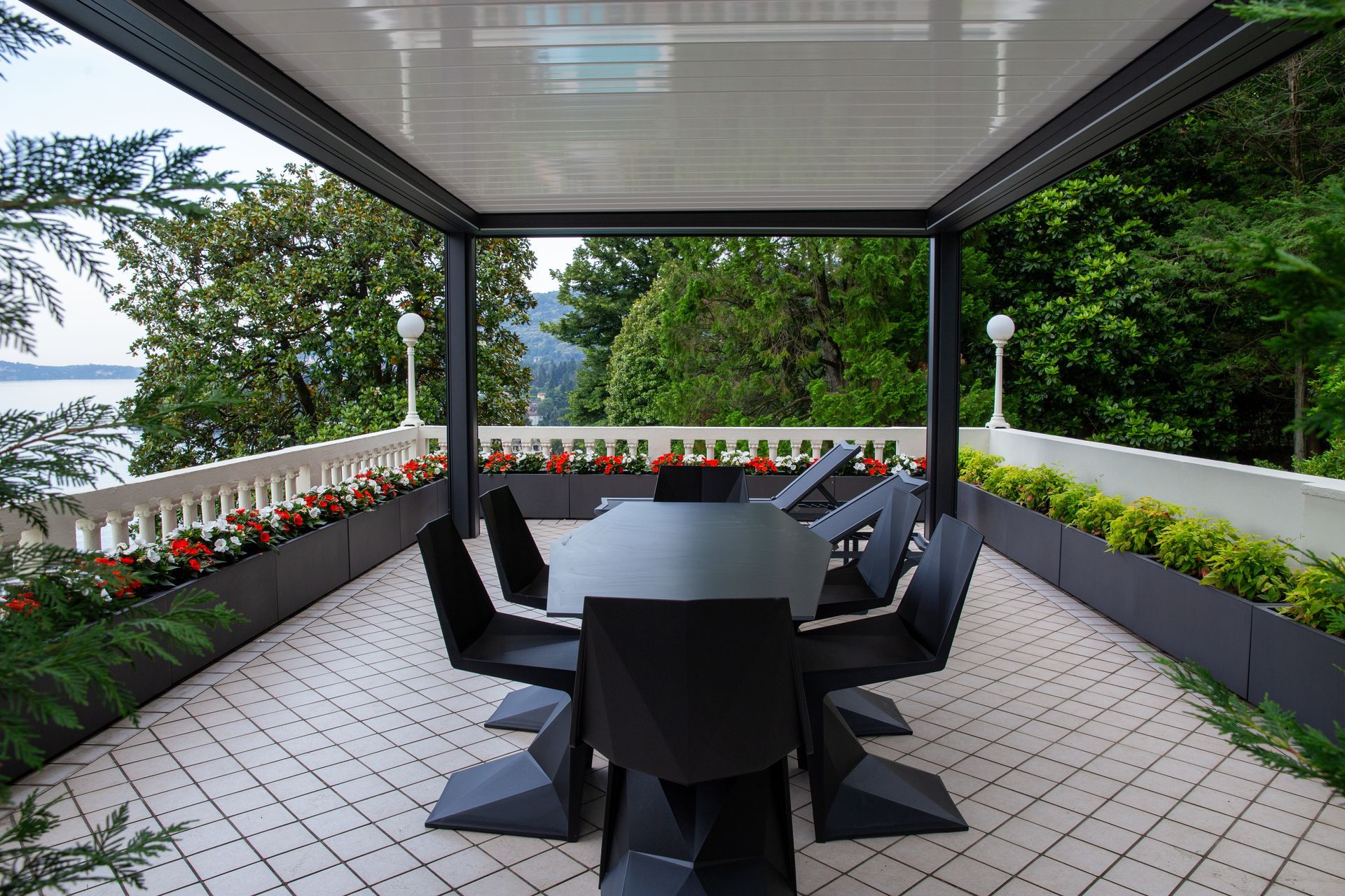 Patio moderno con tavolo da pranzo, sedie nere geometriche e pergola bianca, con vista sugli alberi e su un lago in lontananza.