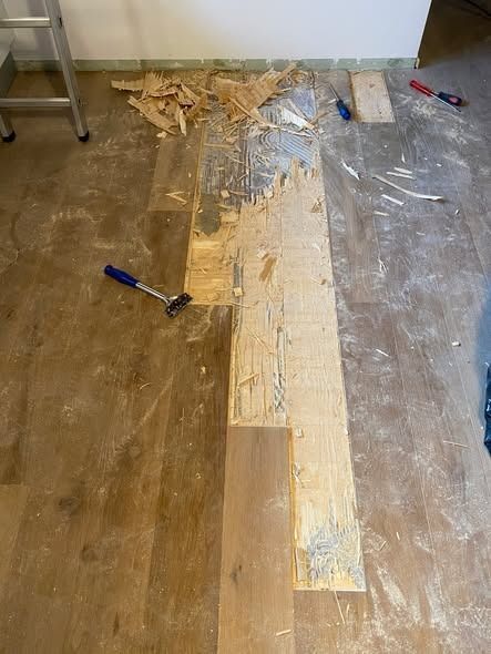 Un pavimento effetto legno parzialmente rimosso, che lascia intravedere il sottofondo, con attrezzi come martello e scalpello sparsi nelle vicinanze.