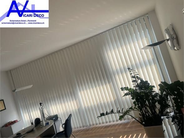 Delle tende verticali bianche coprono un'ampia finestra dell'ufficio, con una pianta in primo piano e una scrivania sullo sfondo.