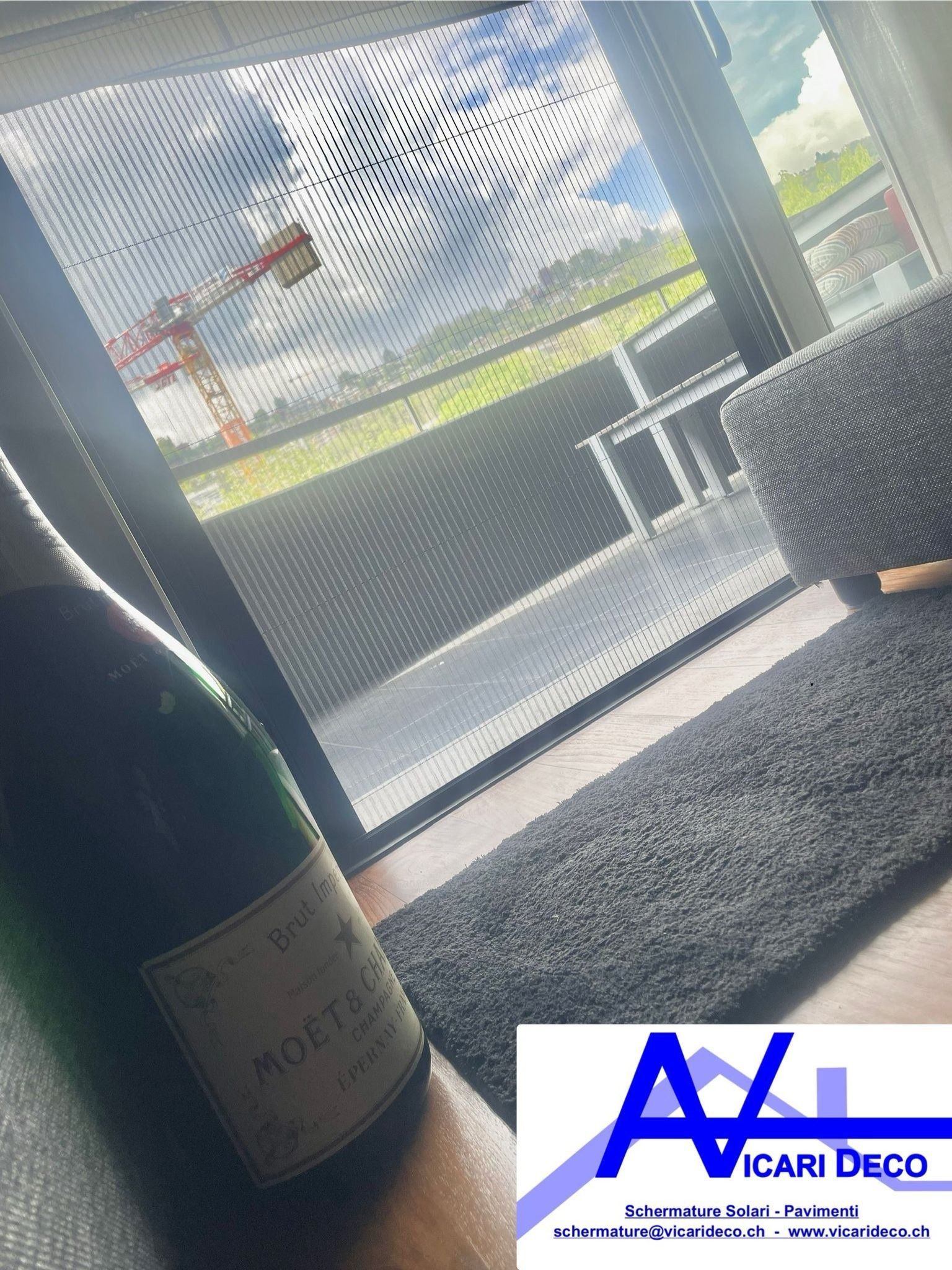 Una bottiglia di champagne Moët & Chandon è appoggiata vicino a una finestra con persiane nere, che si affaccia su un balcone e su una gru da cantiere.