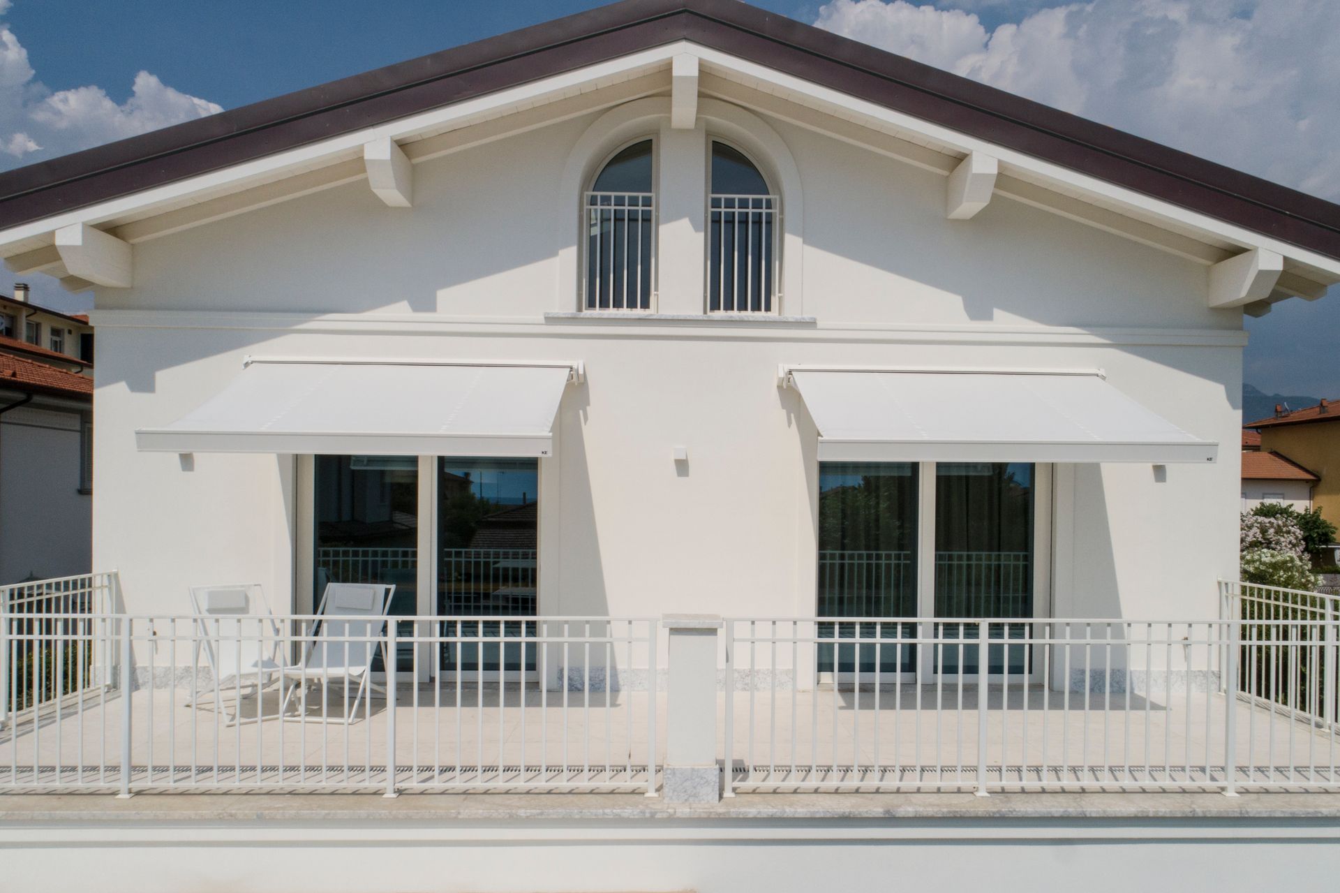 Una casa bianca a due piani con due grandi porte scorrevoli in vetro, tende da sole bianche retrattili e una ringhiera metallica per il balcone.