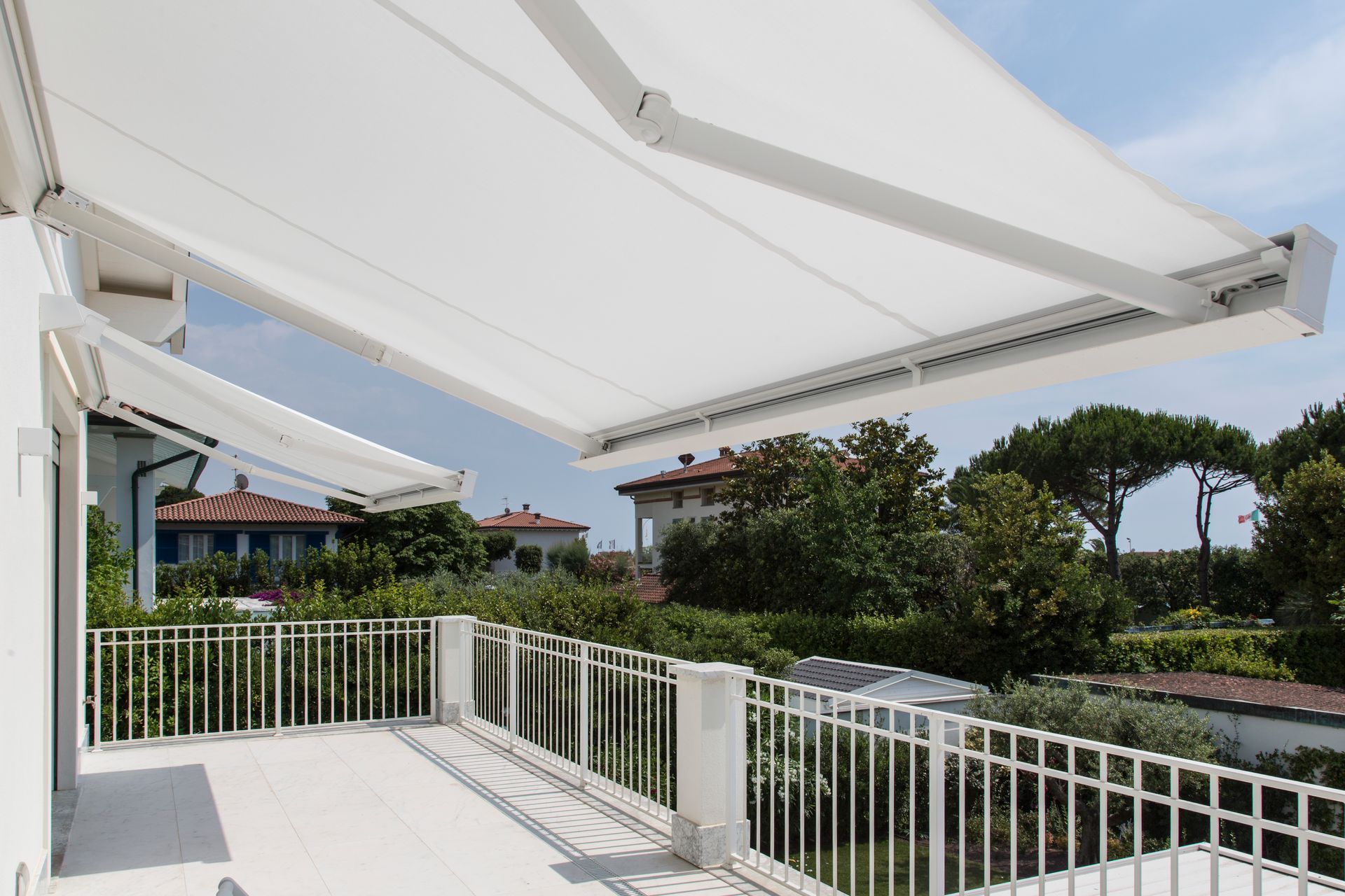 Una tenda da sole retrattile bianca si estendeva su un balcone soleggiato con una ringhiera bianca e una vista su alberi e case.