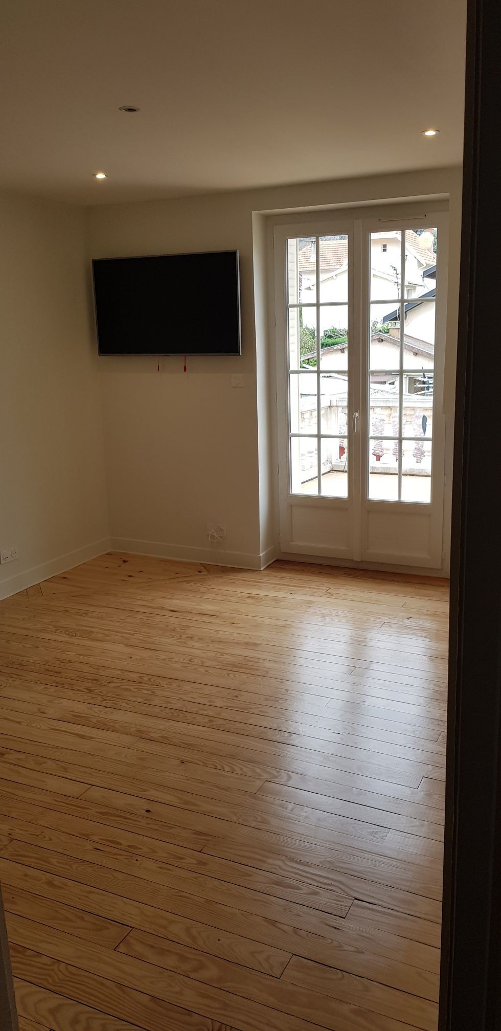 Pièce vide avec parquet, télévision murale et double porte vitrée donnant sur l'extérieur.
