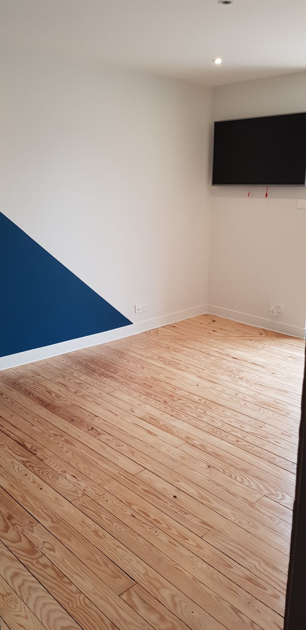 Pièce vide avec parquet clair, murs blancs, un triangle décoratif bleu et une télévision fixée au mur.