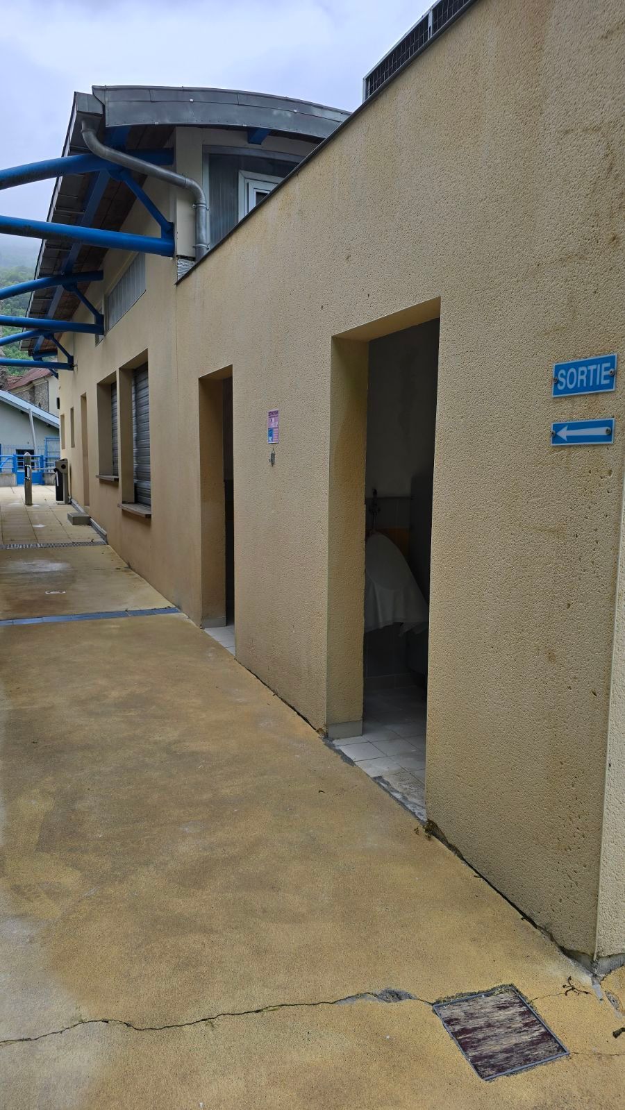 Étroit couloir extérieur jouxtant un bâtiment beige avec plusieurs portes ouvertes et des auvents bleus au-dessus.