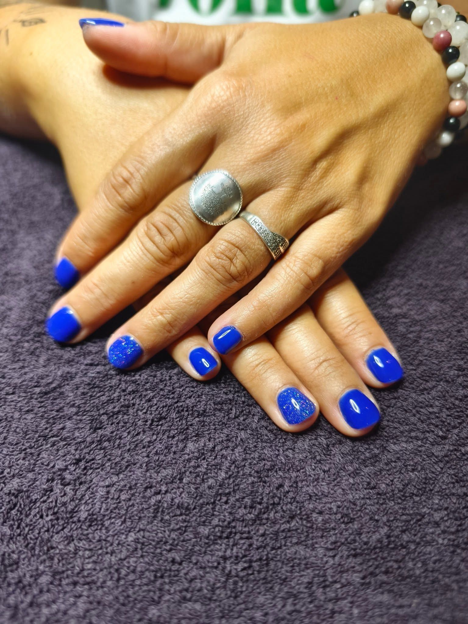 Des mains aux ongles vernis de bleu vif pailleté, ornées de bagues argentées, posées sur une serviette foncée.
