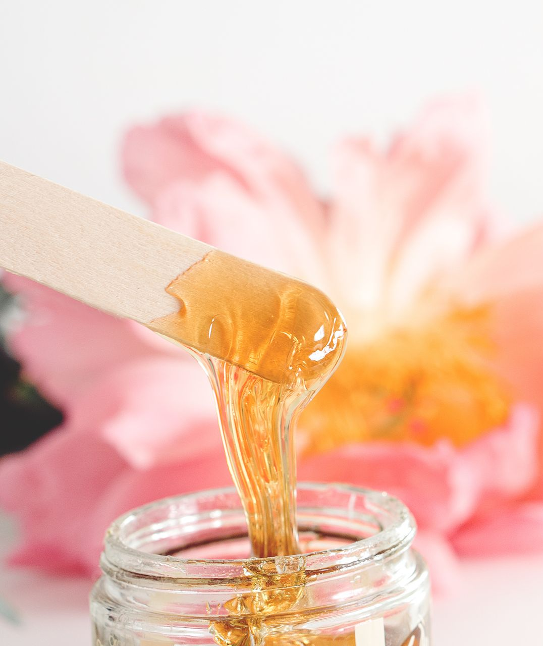 Une spatule soulevant de la cire dorée d’un pot, avec des fleurs roses floues en arrière-plan.