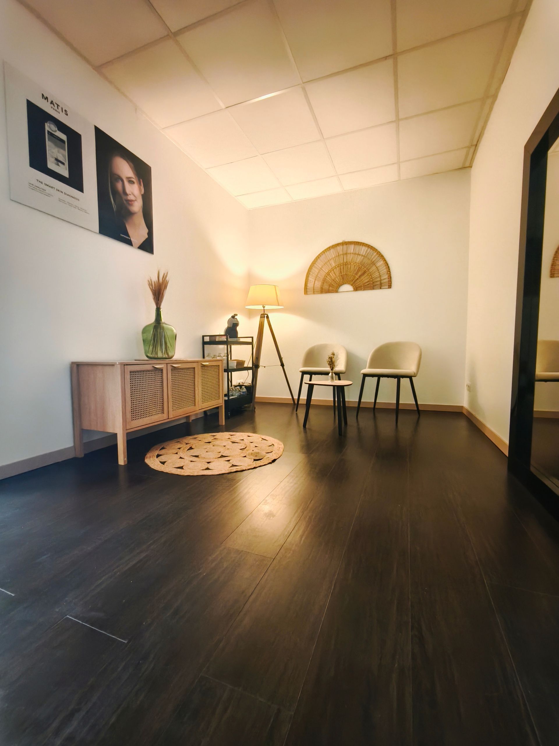 Salle d’attente chaleureuse avec un parquet foncé, deux chaises, une lampe de table, des plantes, des œuvres d’art murales et un éventail décoratif accroché au mur.