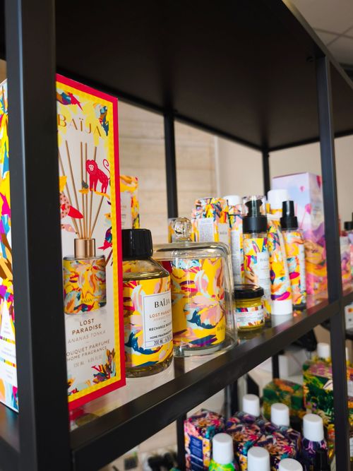 Étagère de bougies colorées et de pots ornés d’étiquettes florales, dans une vitrine de magasin.