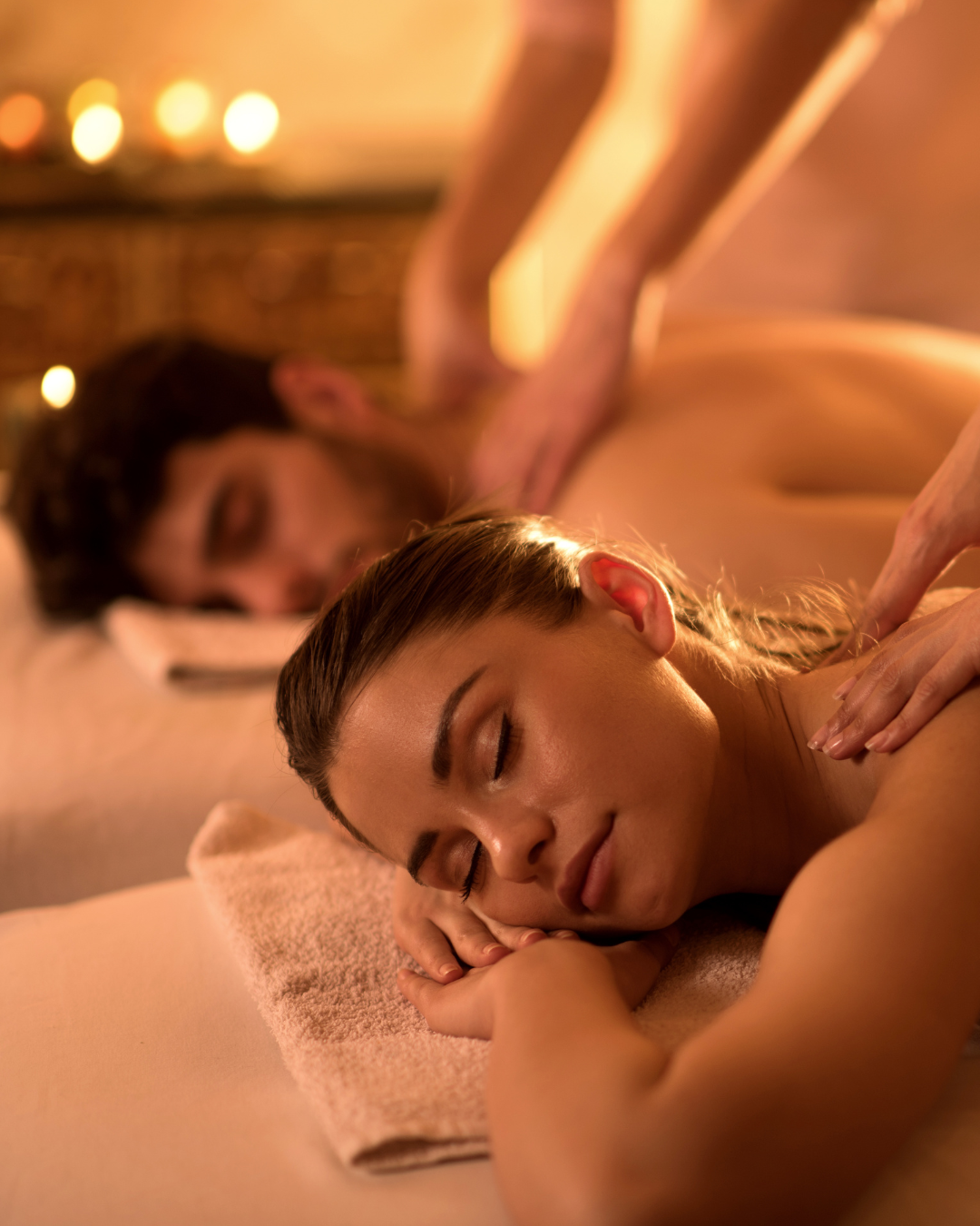 Personne recevant un massage dans un spa, les mains de la masseuse posées sur ses épaules, dans une pièce chaleureuse éclairée à la bougie.