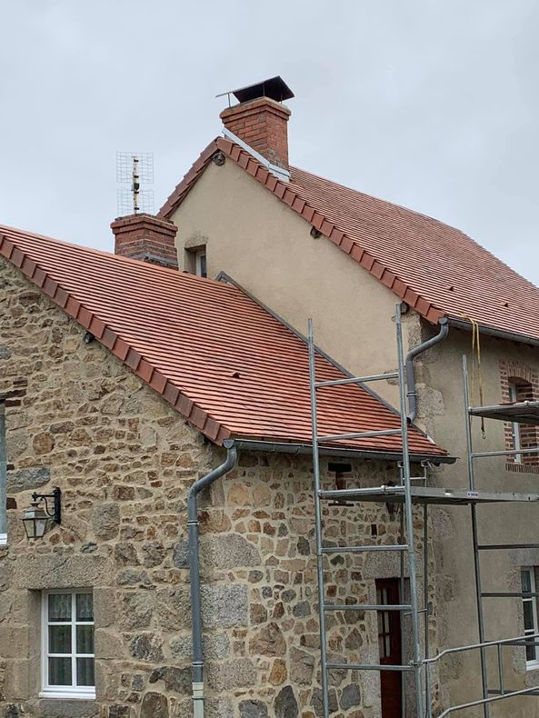 Toits de bâtiments aux tuiles courbes rouge-orange, cheminées en briques, murs en pierre et ciel nuageux.