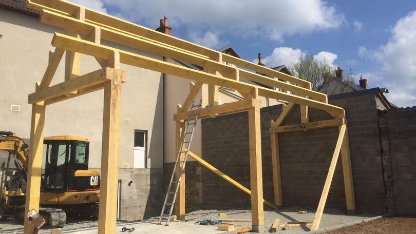Structure à ossature en bois d'un abri d'auto en cours de construction, avec une petite excavatrice garée à proximité.