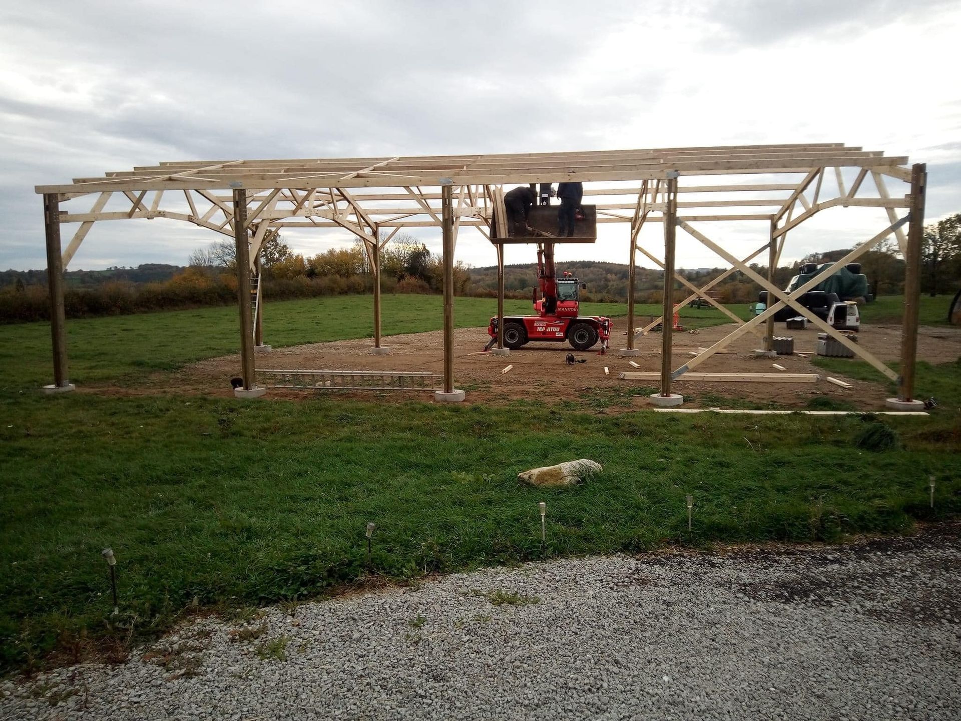 Une structure en bois en construction dans un champ herbeux, avec un chariot élévateur et des ouvriers présents.