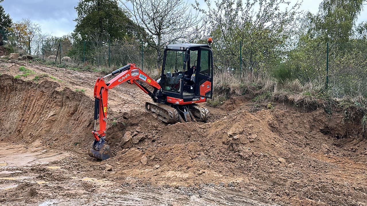 Excavatrice jaune garée sur du gravier et de l'herbe près des arbres sous un ciel bleu.