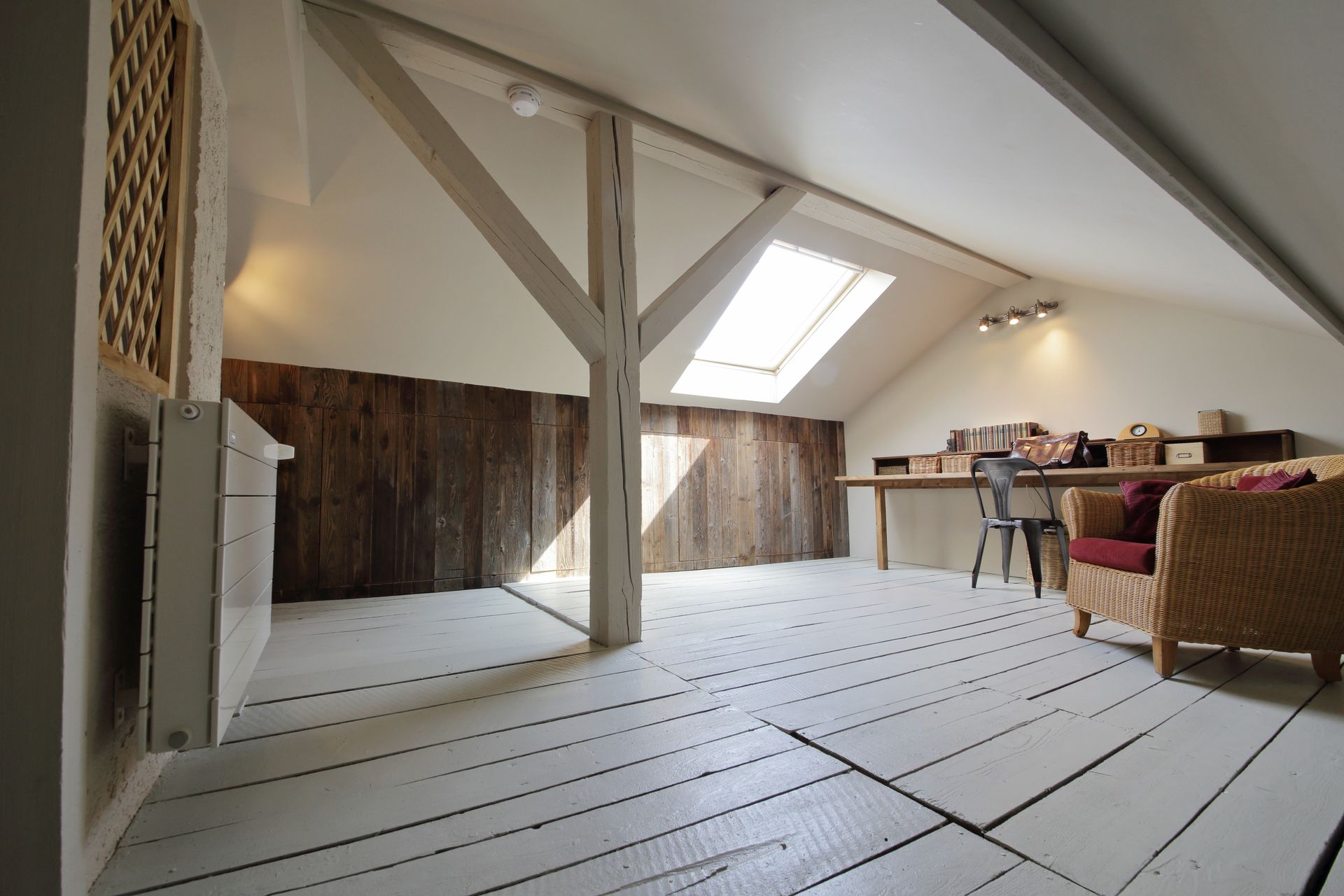 Chambre mansardée avec parquet peint en blanc, poutres en bois, puits de lumière, bureau et fauteuil en osier.