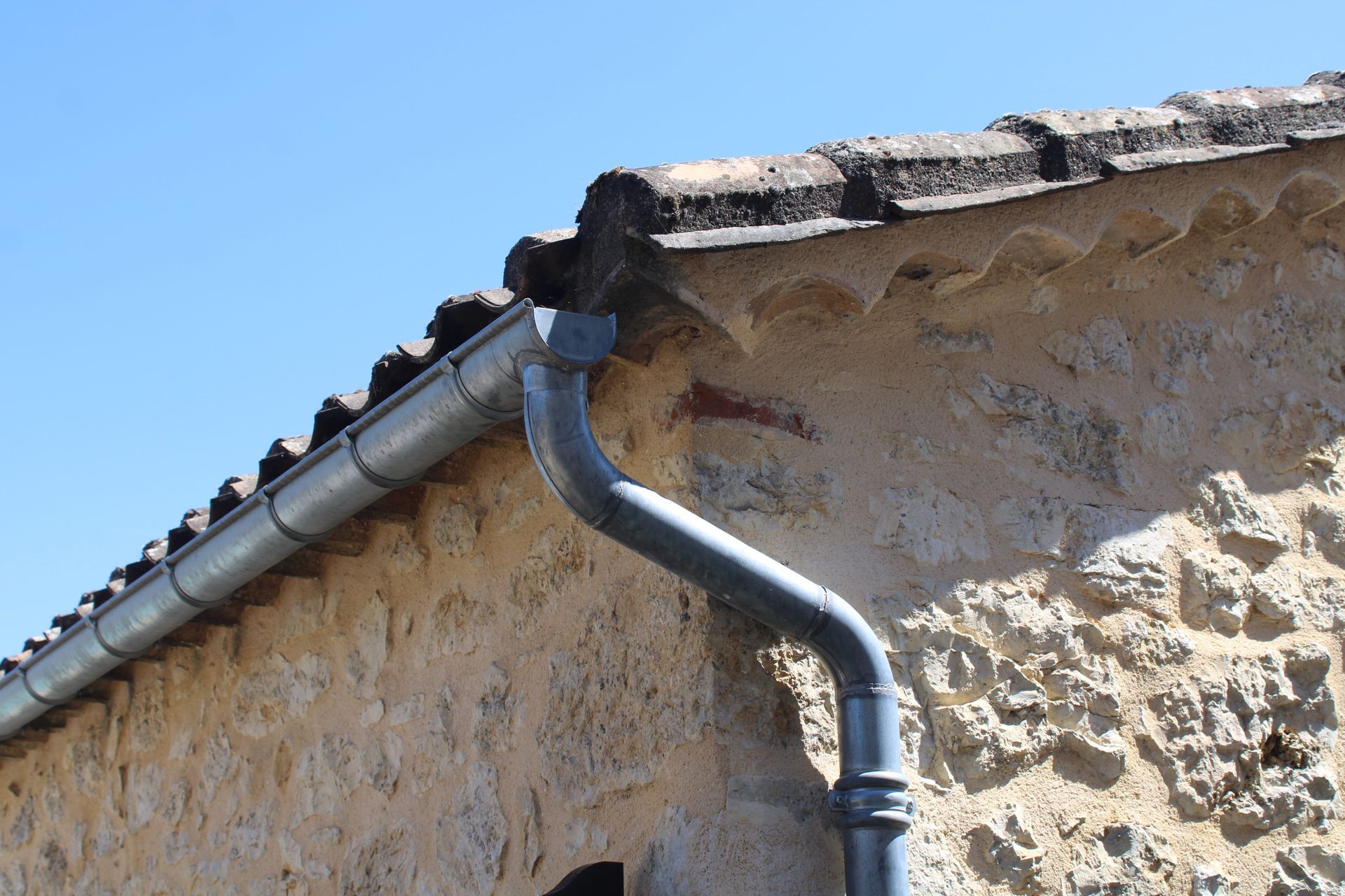 Gouttière en métal gris le long du bord d'un bâtiment en pierre sous un ciel bleu clair.
