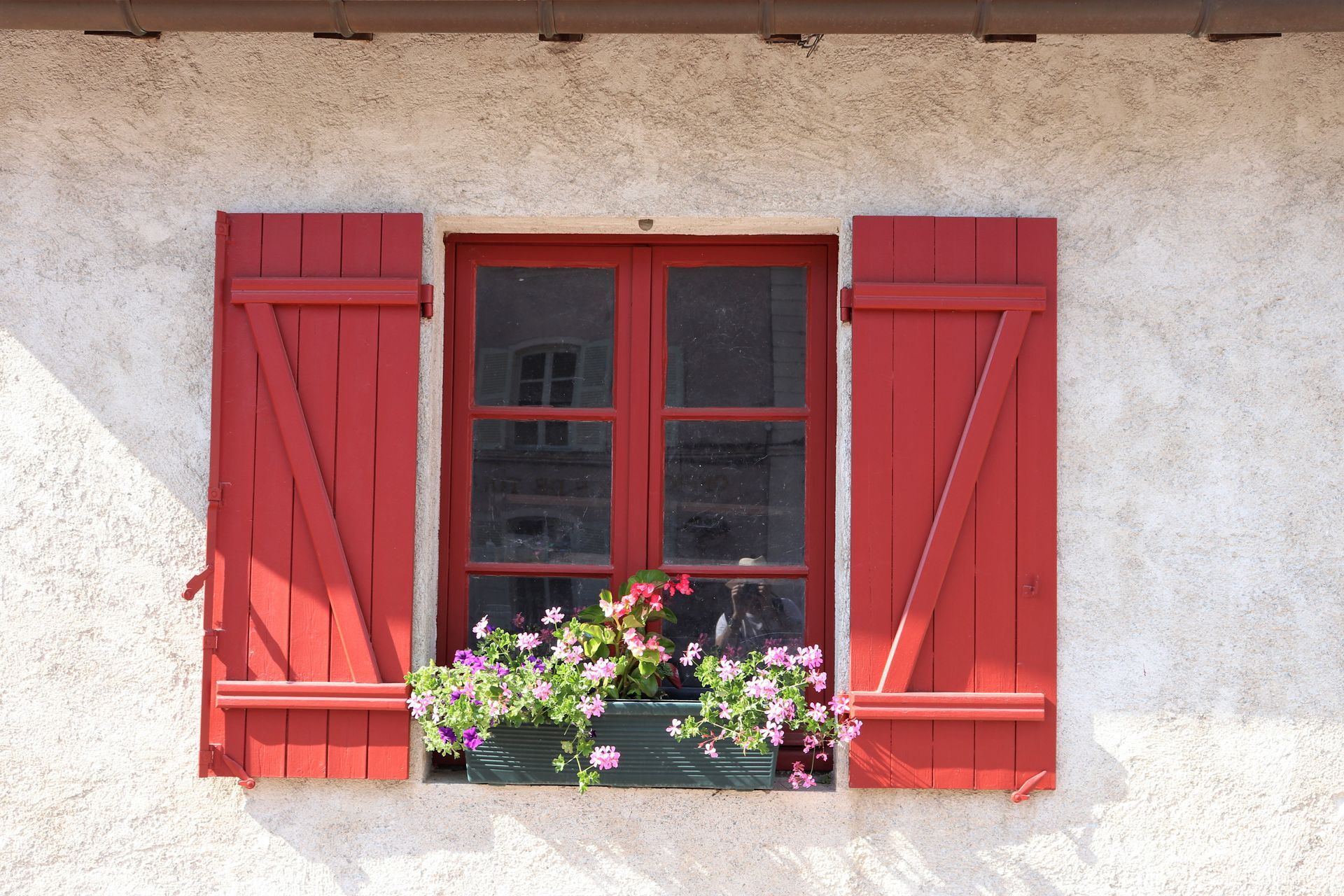 Volets rouges encadrant une fenêtre avec une jardinière de fleurs, sur un mur en stuc beige.