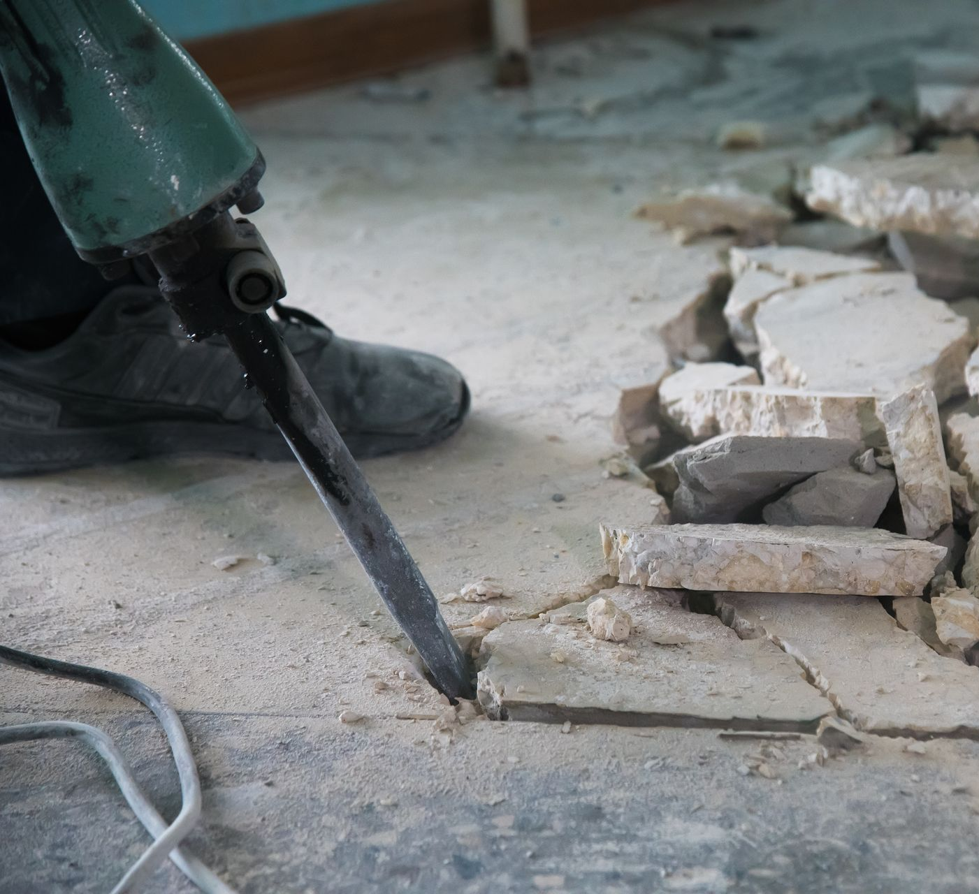 Une personne utilise un marteau-piqueur pour casser le revêtement de sol. Des débris et un outil sont visibles.