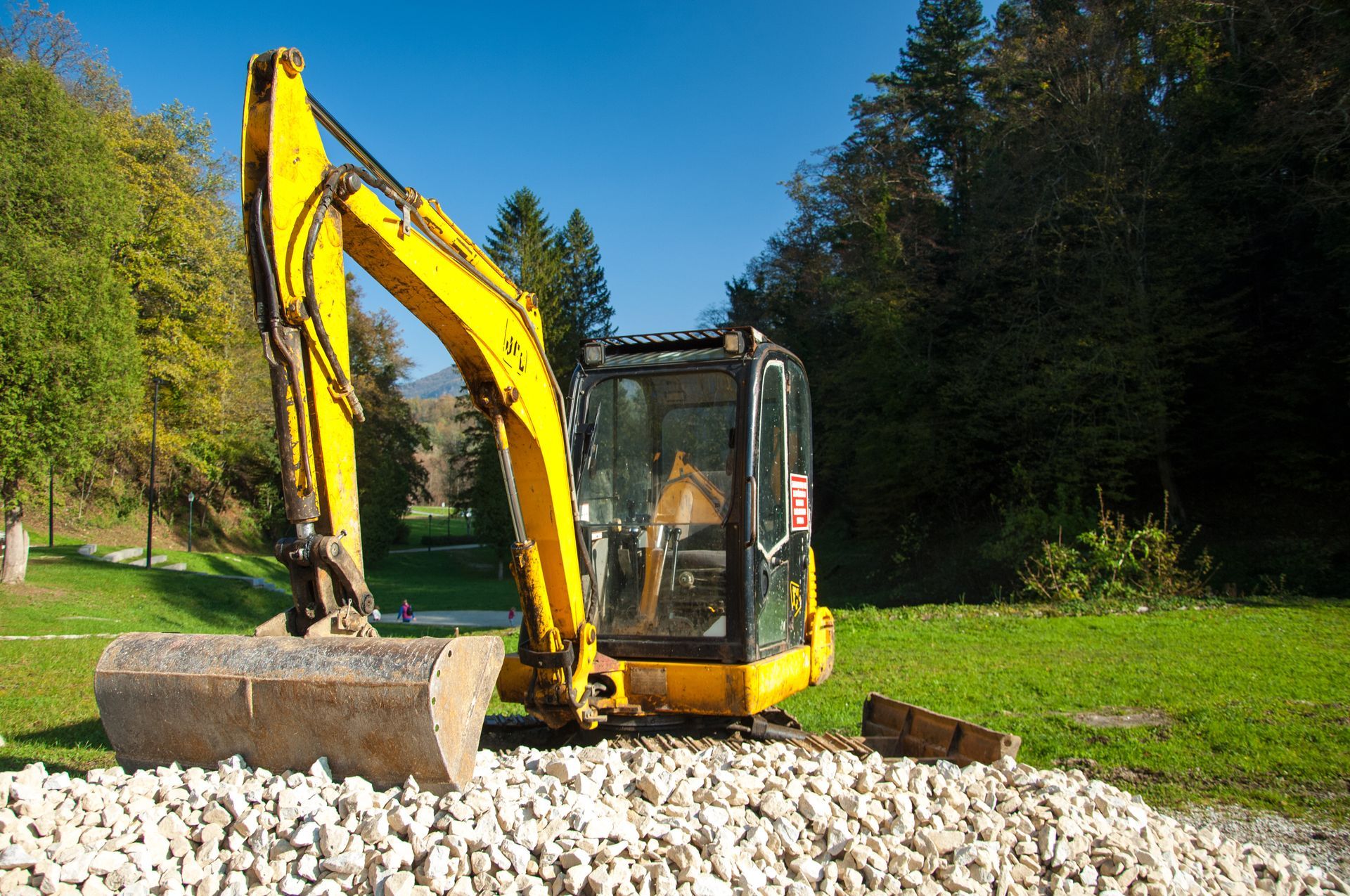 Excavatrice jaune garée sur du gravier et de l'herbe verte près des arbres sous un ciel bleu.