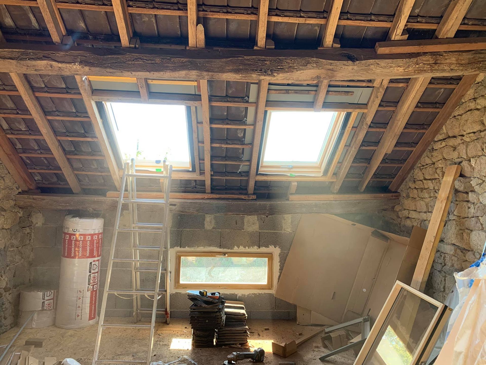 Intérieur d'une pièce en construction avec deux puits de lumière, des poutres apparentes et une petite fenêtre.