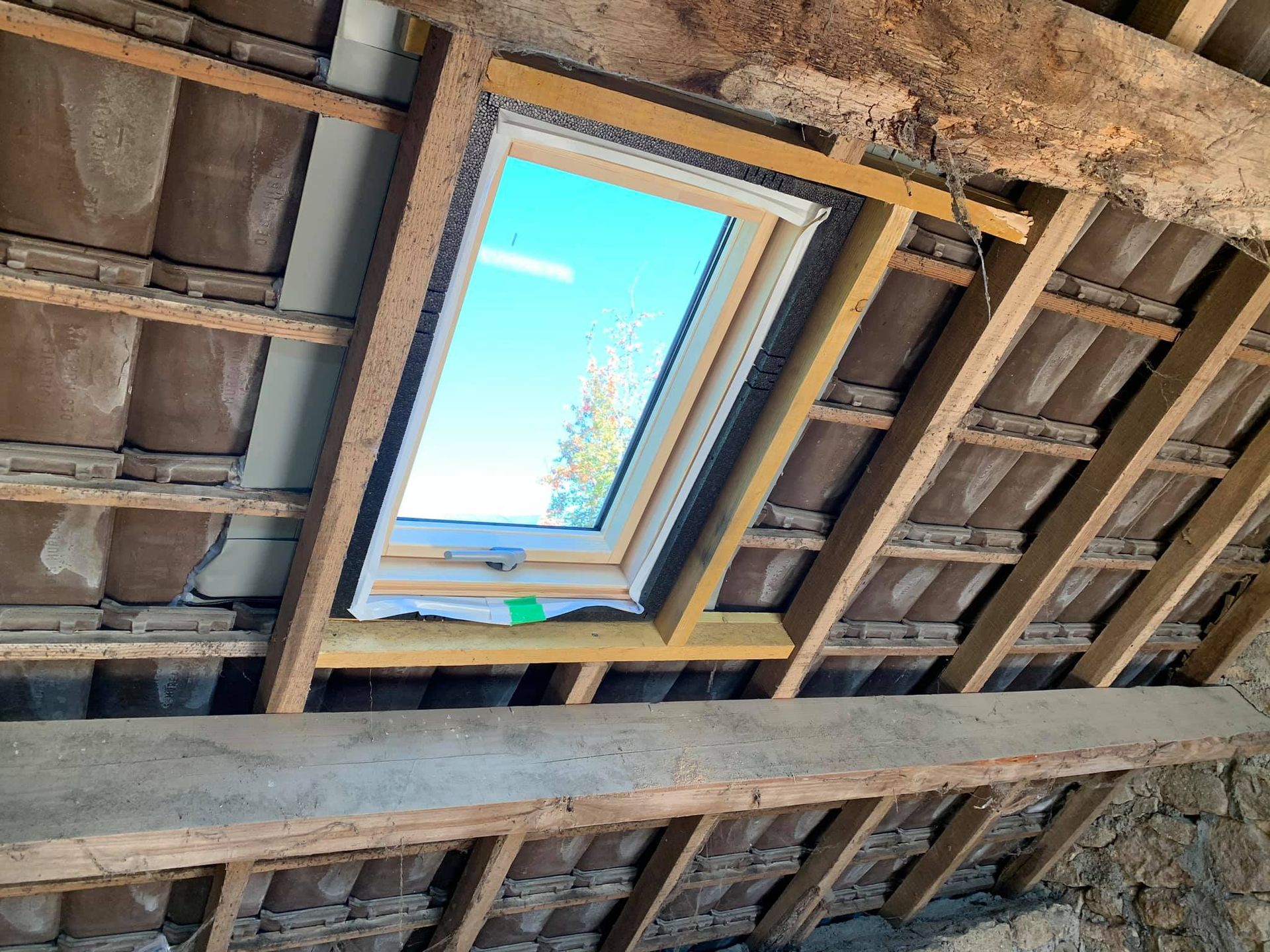 Puits de lumière installé dans un toit mansardé, encadré de bois, ciel bleu visible.