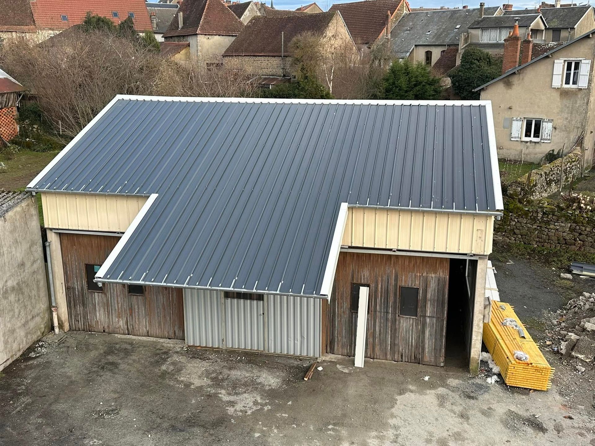 Un petit bâtiment en bois avec un toit en métal gris foncé, de vieilles portes en bois et des bâtiments environnants.