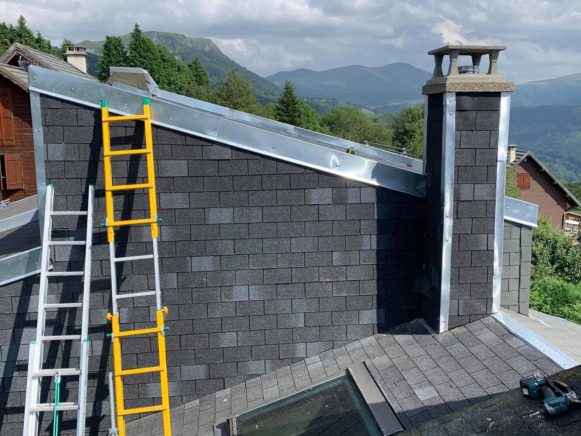 Toiture en bardeaux foncés avec cheminée. Des échelles sont posées sur le toit. Montagnes en arrière-plan.