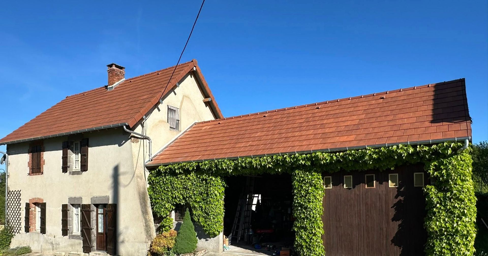 Maison en pierre avec toit en tuiles rouges, garage attenant couvert de vignes vertes, sous un ciel bleu.