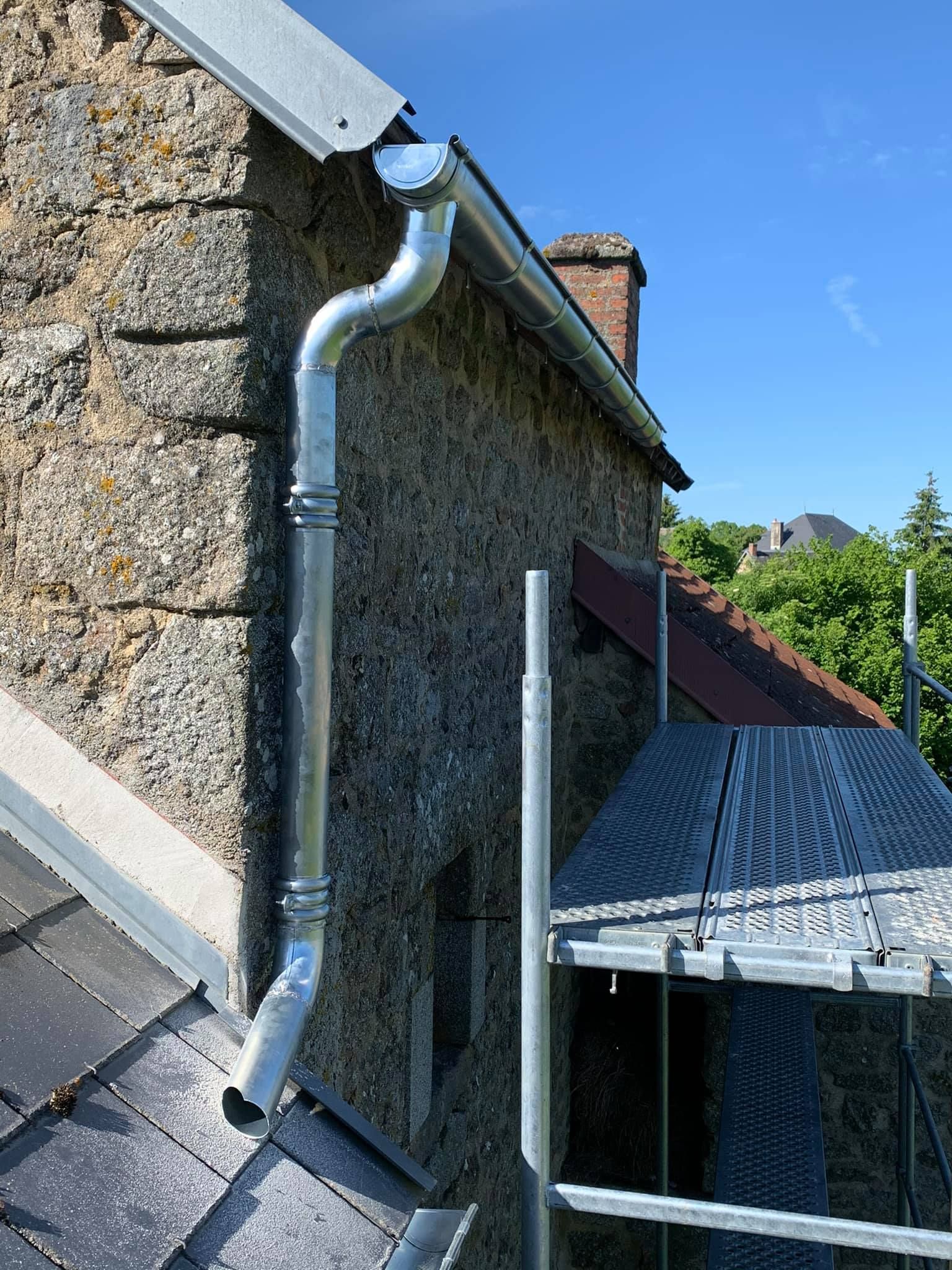 Gouttière et descente pluviale métalliques sur un bâtiment en pierre, vue d'angle. Un échafaudage est partiellement visible en dessous.