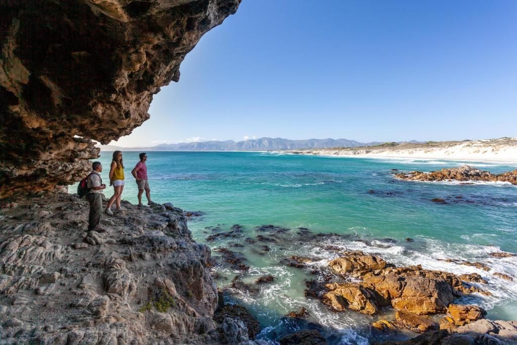 Three people stand near a rocky shoreline under a cave, overlooking turquoise ocean and white sand.