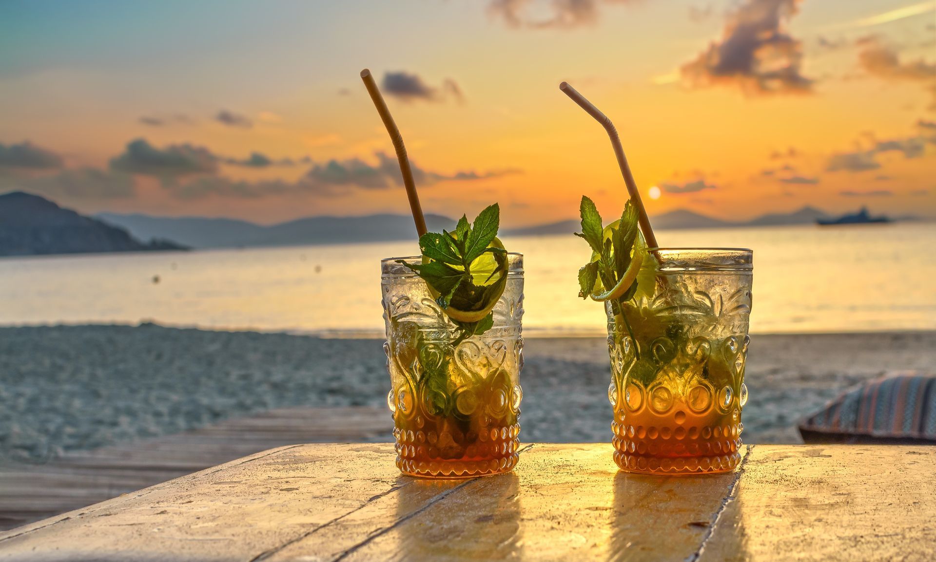 Two cocktails with straws and mint on a table, beach sunset in the background.
