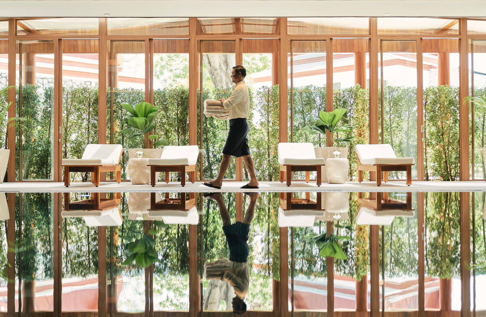 A person walking along a reflective pool, holding items in a spa-like setting with seating and greenery.